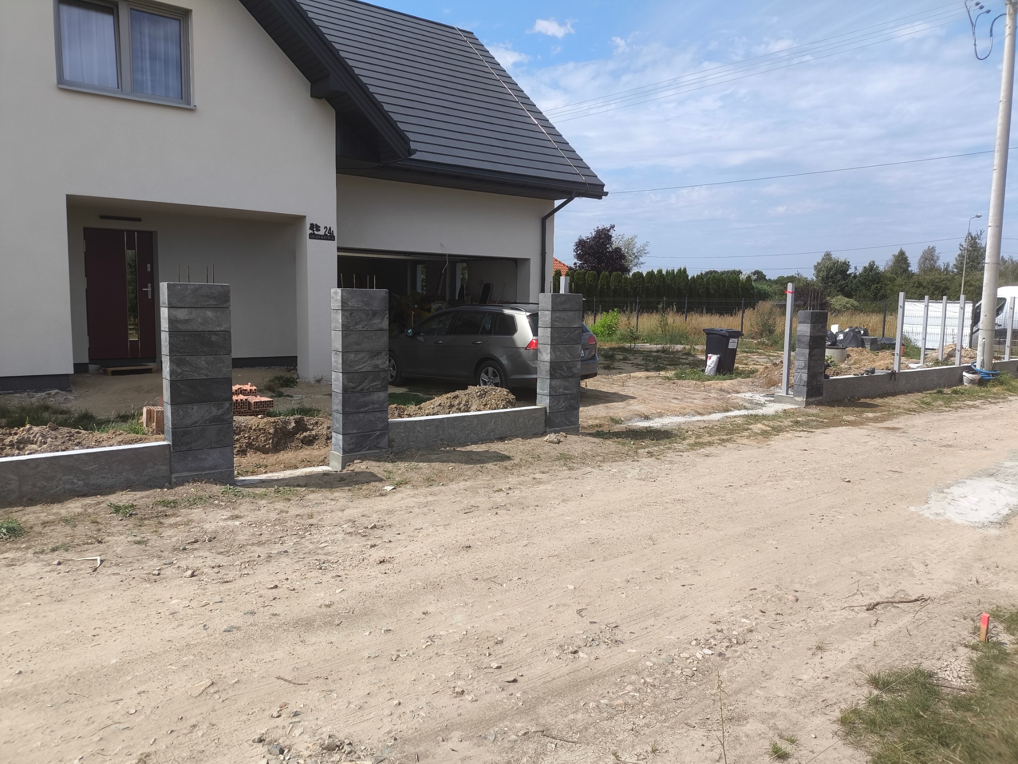 Montaż ogrodzenia z szarej kostki betonowej przy domu jednorodzinnym. Widoczne słupki i podmurówka w trakcie budowy. W tle zaparkowany samochód.