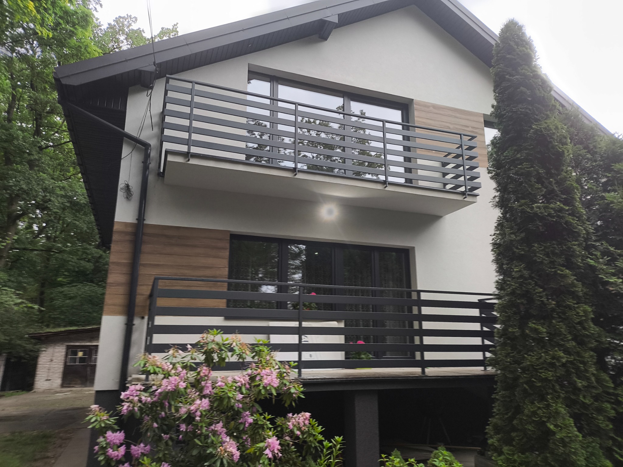 Dwukondygnacyjny dom z balkonem i nowoczesną balustradą z poziomych, czarnych profili. Elewacja w odcieniach bieli i drewna, otoczona zielenią.