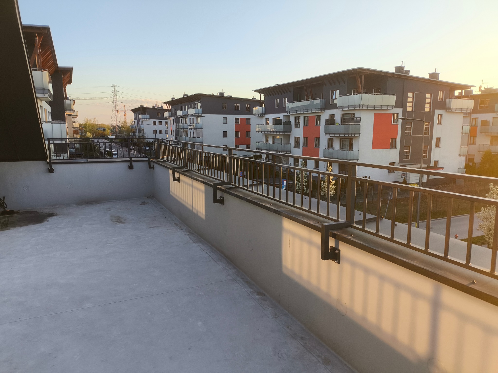 Nowoczesna, metalowa balustrada balkonowa w nowym budownictwie, z widokiem na osiedle i słup wysokiego napięcia w tle, oświetlona ciepłym światłem zachodzącego słońca.