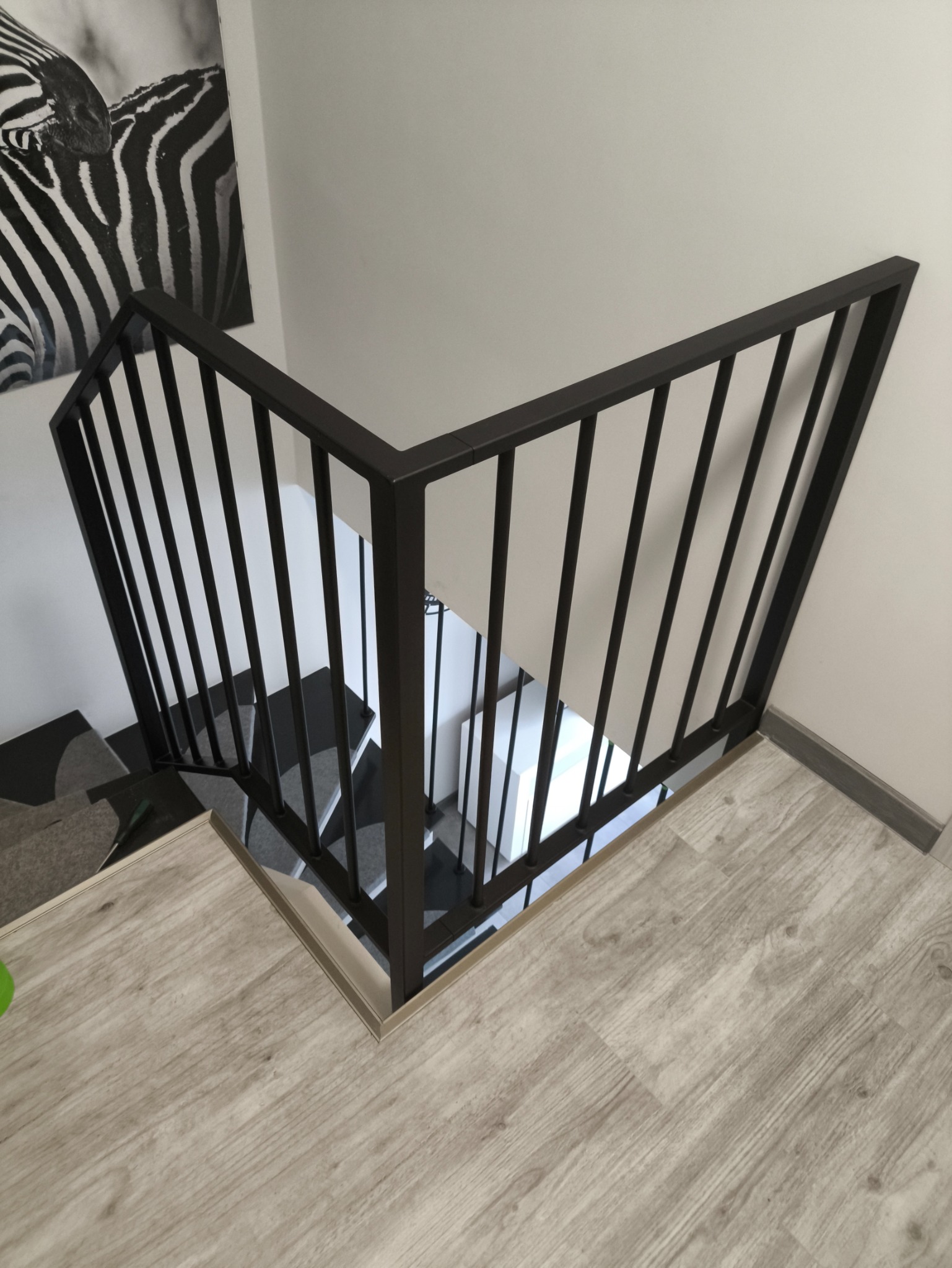Nowoczesna, czarna balustrada schodowa z pionowych prętów, widoczna z góry, z drewnianymi panelami podłogowymi i dekoracją zebrą na ścianie.