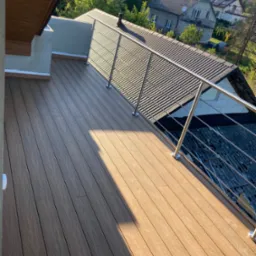 Nowoczesny taras na dachu z desek kompozytowych w odcieniach brązu, zabezpieczony metalową balustradą, widok na okoliczne domy i zieleń.