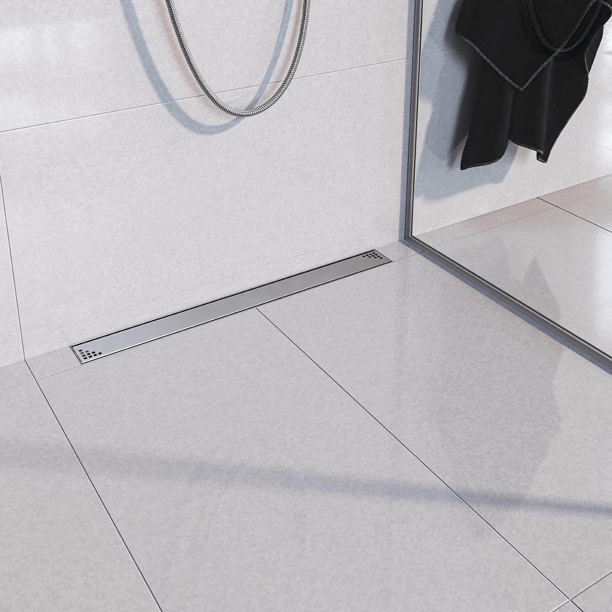 Nowoczesna łazienka z odpływem liniowym w podłodze wyłożonej jasnymi, kwadratowymi płytkami, widoczny fragment kabiny prysznicowej i wąż prysznicowy.