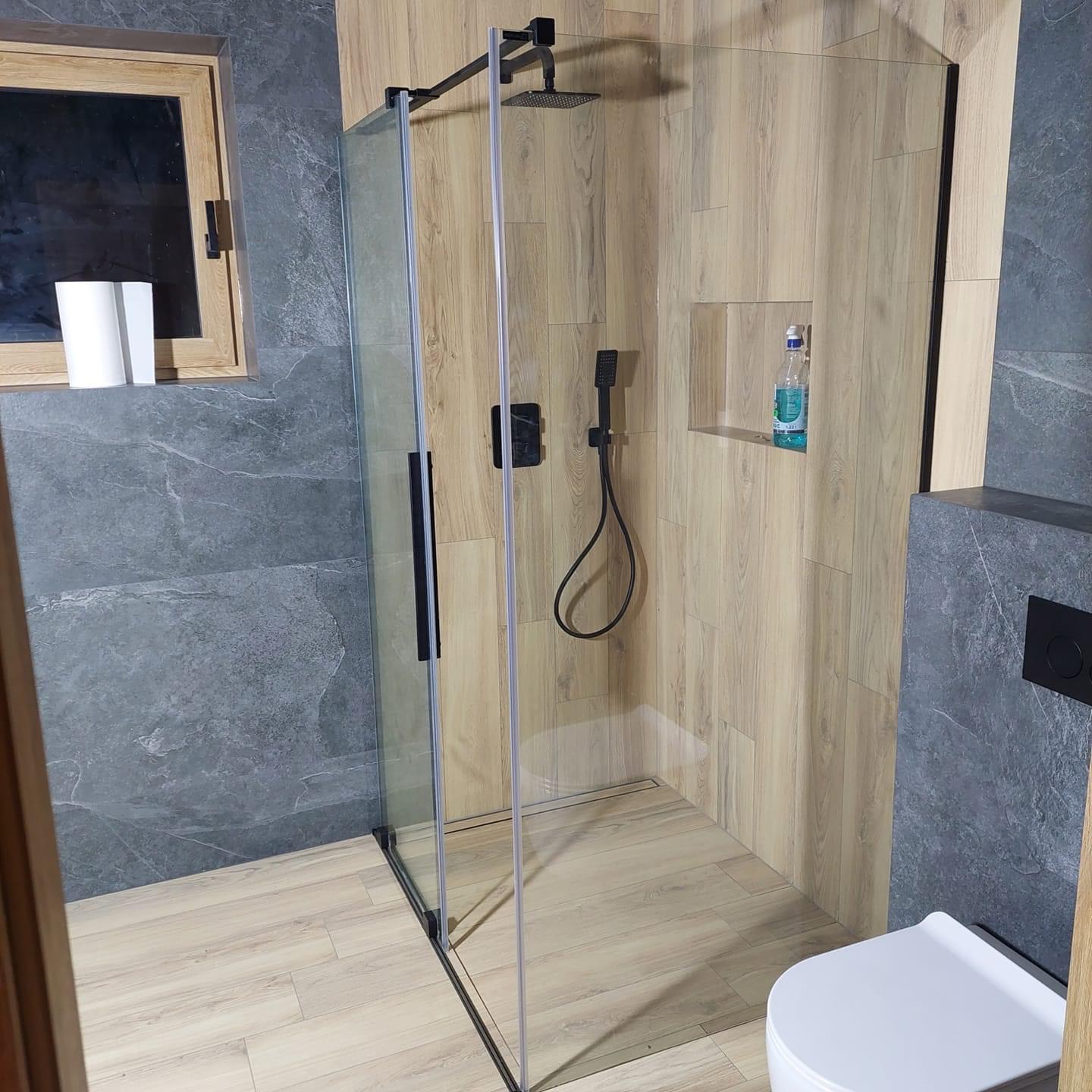 Nowoczesna kabina prysznicowa z czarną armaturą i drewnopodobnymi płytkami, wnęka z płynem do mycia, szare płytki na ścianie obok okna.