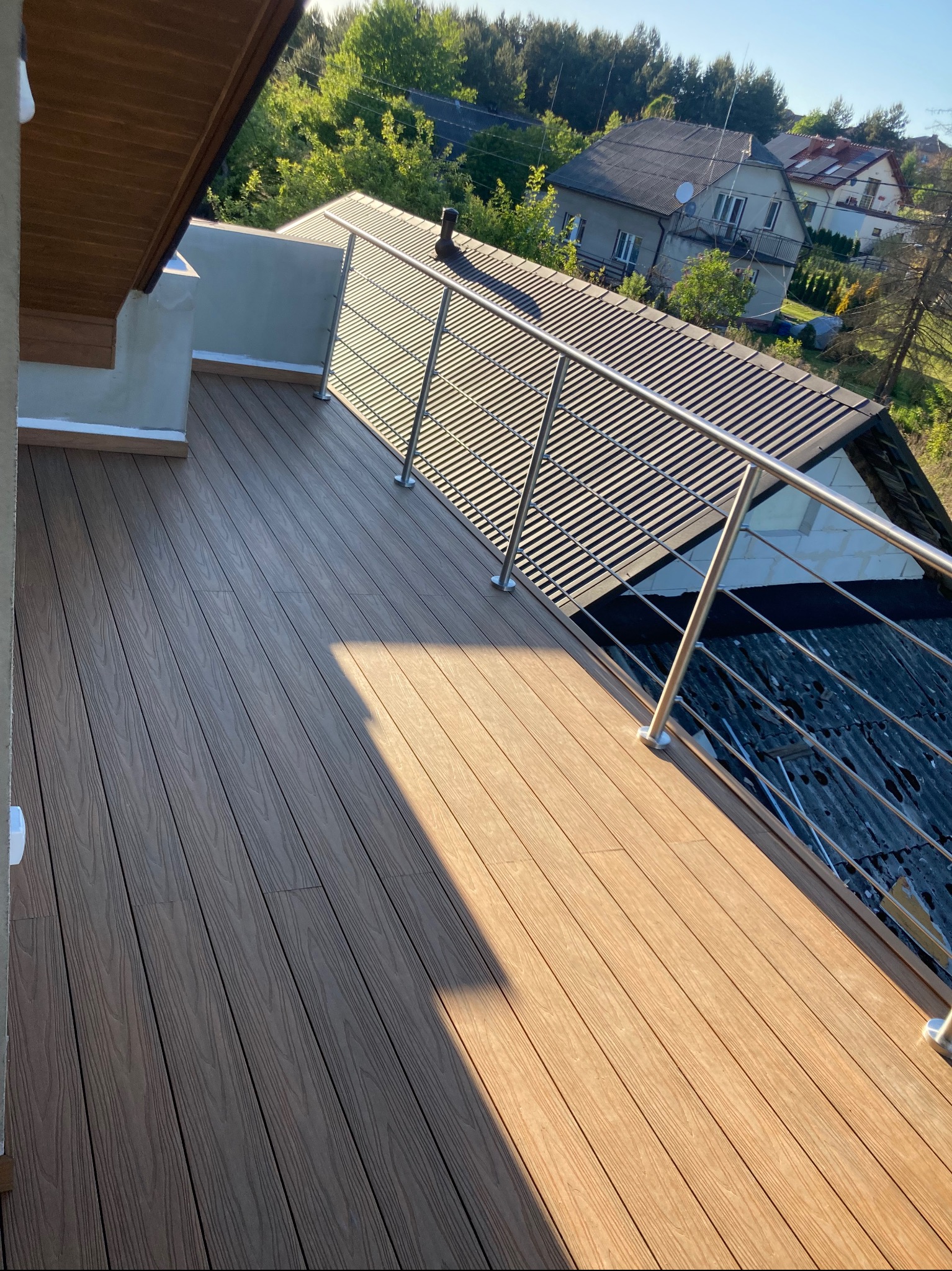 Nowoczesny taras na dachu z desek kompozytowych w odcieniach brązu, zabezpieczony metalową balustradą, widok na okoliczne domy i zieleń.