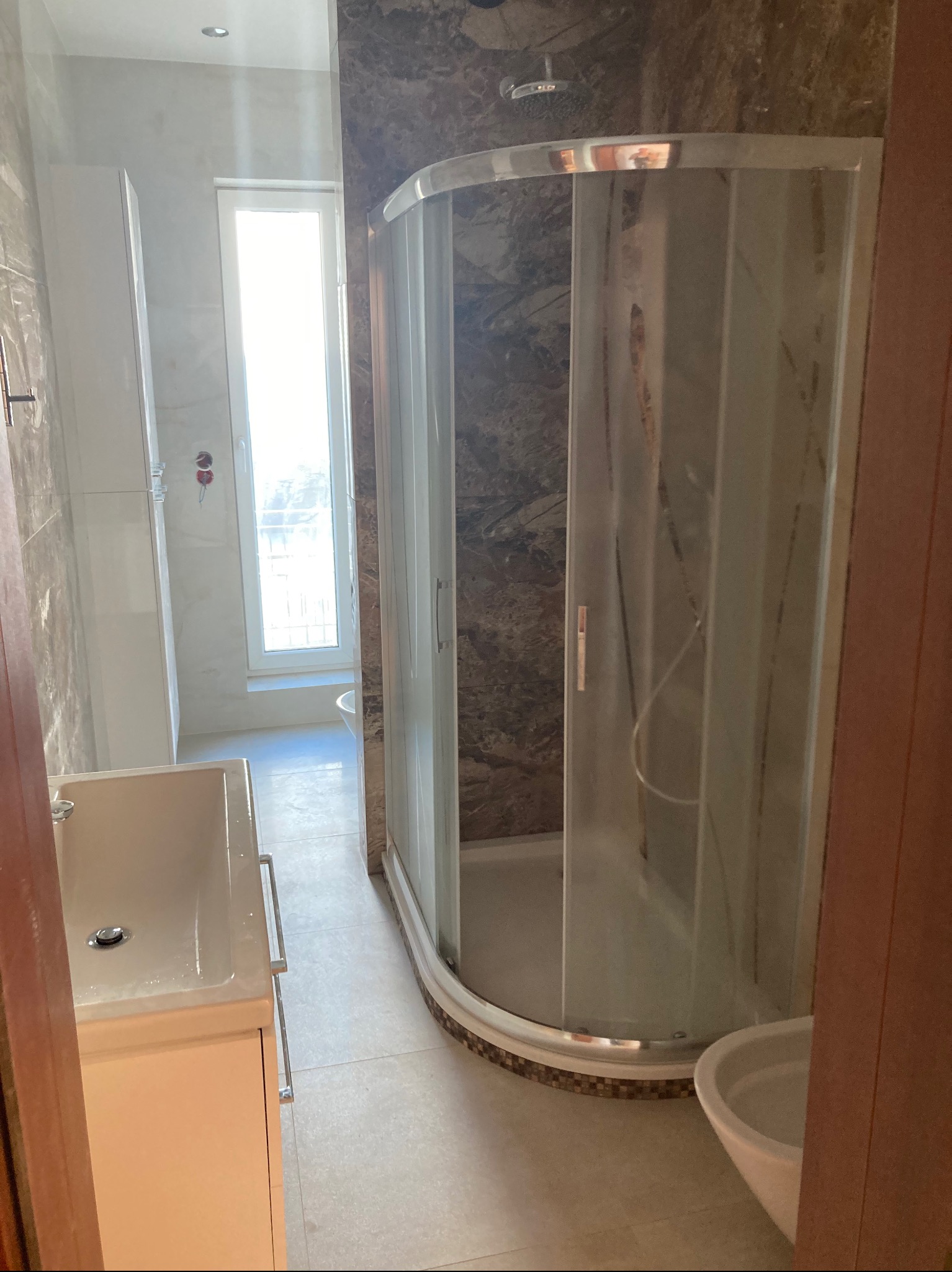 Nowoczesna łazienka z kabiną prysznicową z zaokrąglonymi szybami, umywalką z szafką w kolorze ecru i marmurowymi płytkami na ścianach.