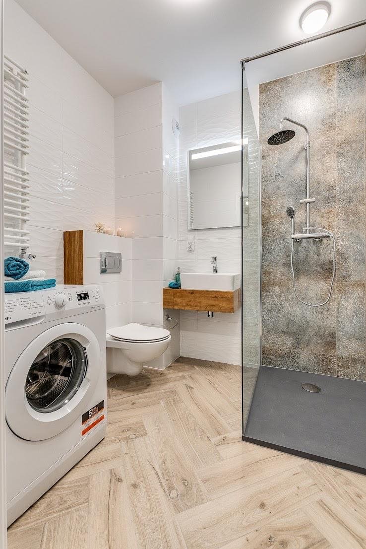Jasna łazienka z pralką, toaletą, umywalką na drewnianej szafce i kabiną prysznicową z deszczownicą, wykończona białymi płytkami i drewnopodobną podłogą ułożoną w jodełkę.