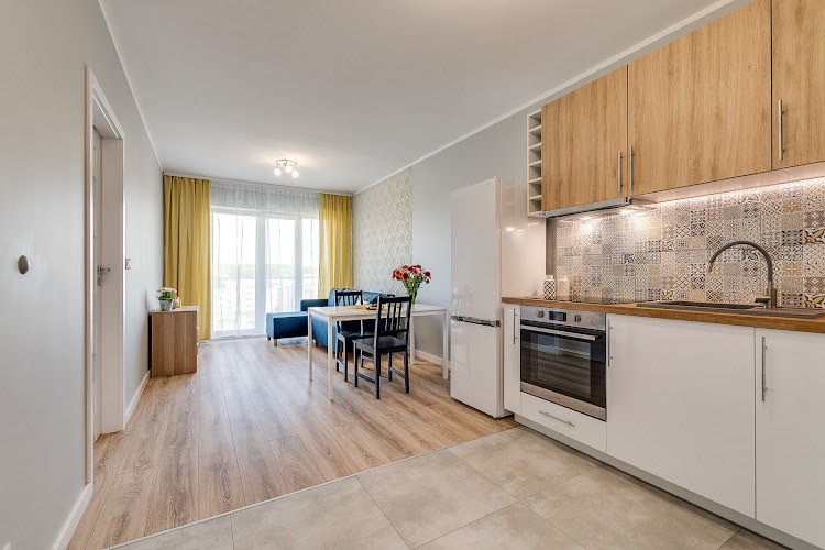 Jasne wnętrze mieszkania z aneksem kuchennym, drewniane panele podłogowe przechodzą w jasne płytki, białe meble kuchenne z drewnianym blatem i ozdobnymi płytkami nad blatem, w tle salon z niebieską...