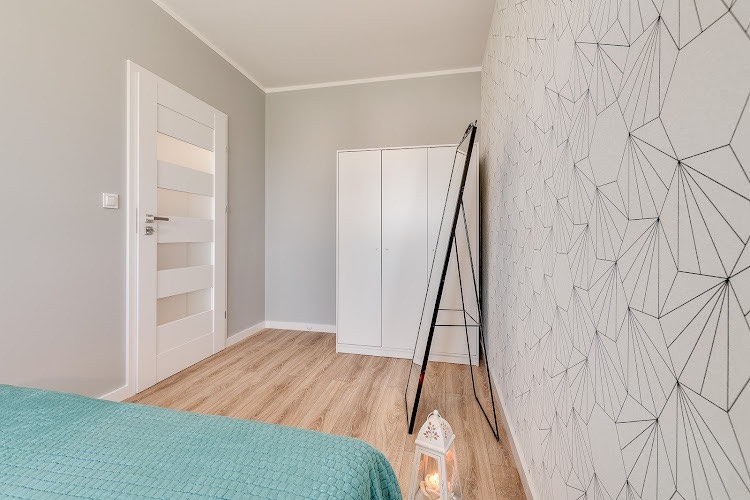 Jasne wnętrze pokoju po remoncie: białe drzwi z przeszkleniami, drewnopodobne panele, biała szafa, lustro oprawione w czarną ramę, ściana z geometrycznym wzorem, dekoracyjna latarenka i fragment...