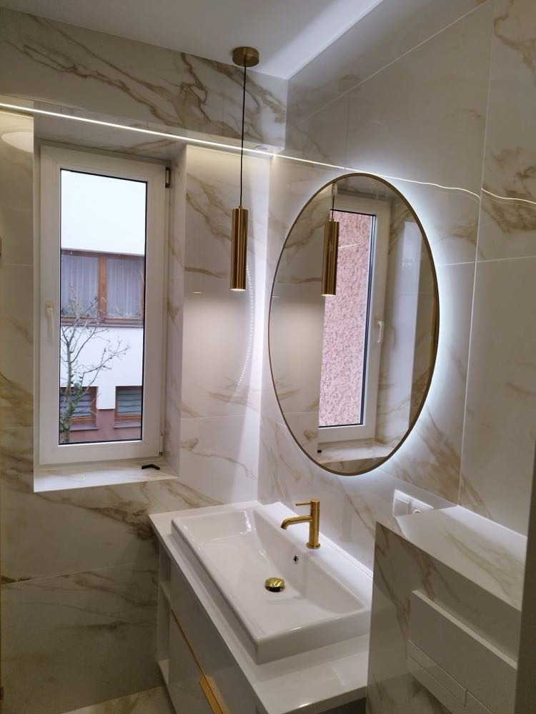 Elegancka łazienka z marmurowymi płytkami, owalnym lustrem z podświetleniem LED, białą umywalką z złotą armaturą i wiszącymi złotymi lampami, widok na okno z widokiem na budynek mieszkalny.