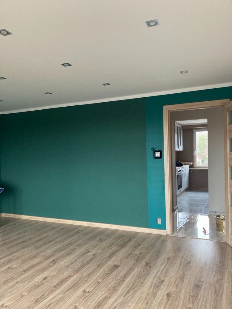 Pomieszczenie po malowaniu ścian na ciemny turkus, widoczne panele podłogowe i fragment kuchni w tle.