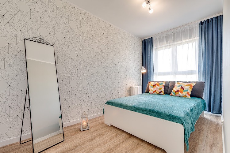 Jasna sypialnia z podwójnym łóżkiem, geometryczną tapetą, niebieskimi zasłonami i wysokim lustrem w czarnej ramie.