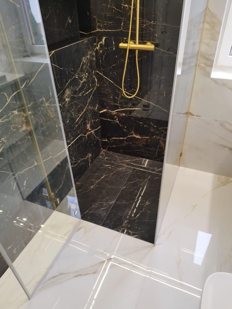 Kabina prysznicowa wyłożona czarnym marmurem ze złotymi żyłami, złota armatura, białe płytki podłogowe z delikatnym marmurkowym wzorem, widok z góry.