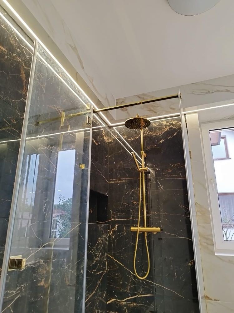 Luksusowy prysznic typu walk-in z czarnymi marmurowymi płytkami, złotą armaturą i subtelnym oświetleniem LED, widok na zewnętrzny krajobraz przez szybę.