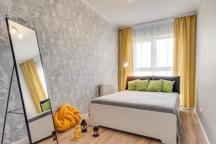 Nowoczesna sypialnia z łóżkiem, żółtymi zasłonami, lustrem w czarnej ramie i tapetą w liście, uzupełniona dekoracyjną poduszką w kształcie węzła i lampionami.