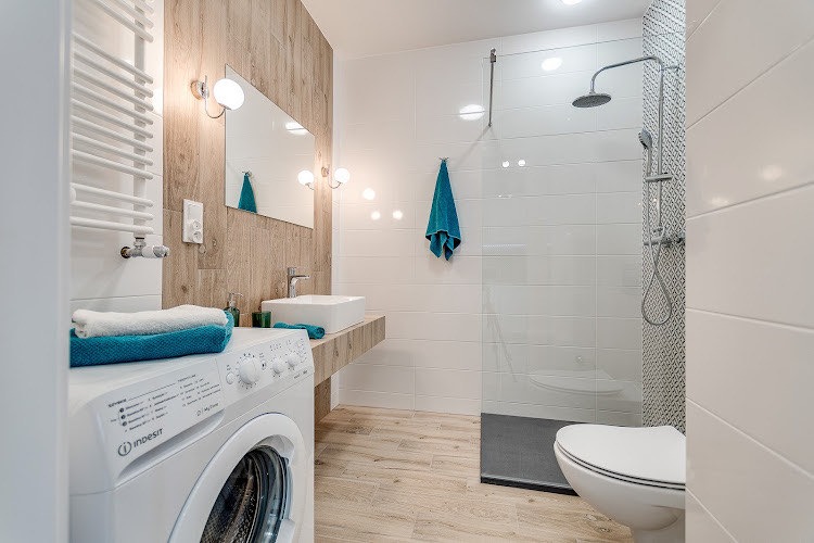 Jasna łazienka z drewnianymi akcentami, kabiną prysznicową typu walk-in, umywalką nablatową i pralką z ułożonymi na niej turkusowymi ręcznikami.