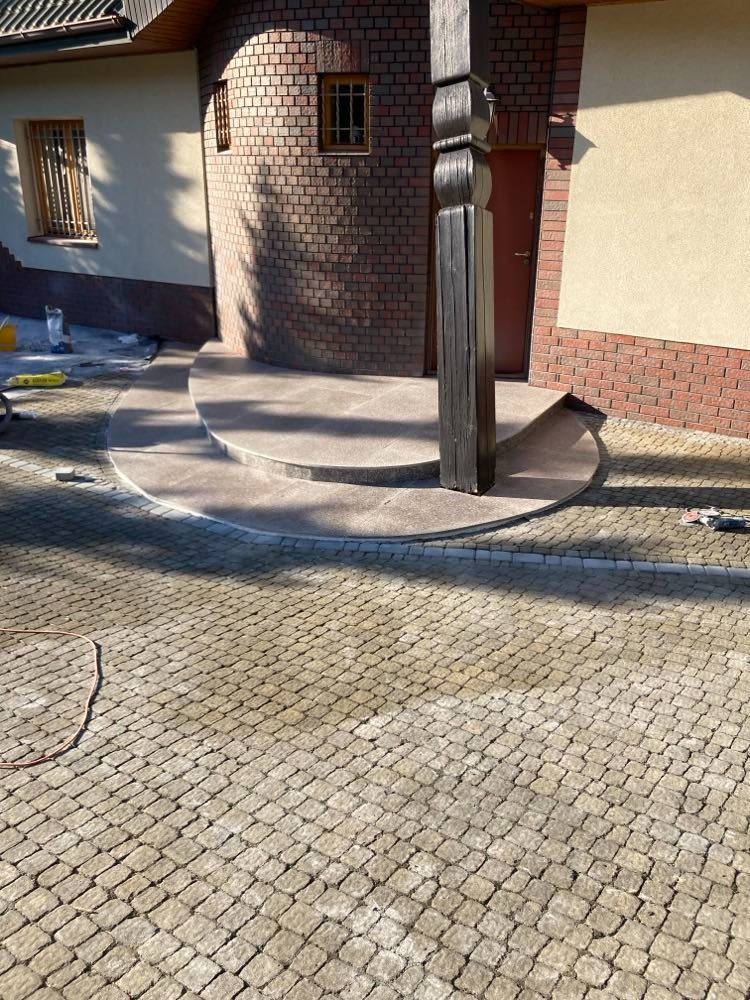 Ułożona kostka brukowa przed wejściem do budynku z elementami wykończenia z cegły, widoczny fragment drewnianej kolumny i schodów.