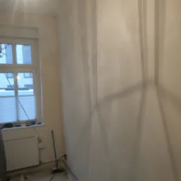 Ściana w pokoju po nałożeniu wstępnej warstwy gładzi, widoczne nier&oacute;wności i ślady szpachlowania, okno z żaluzjami w tle.