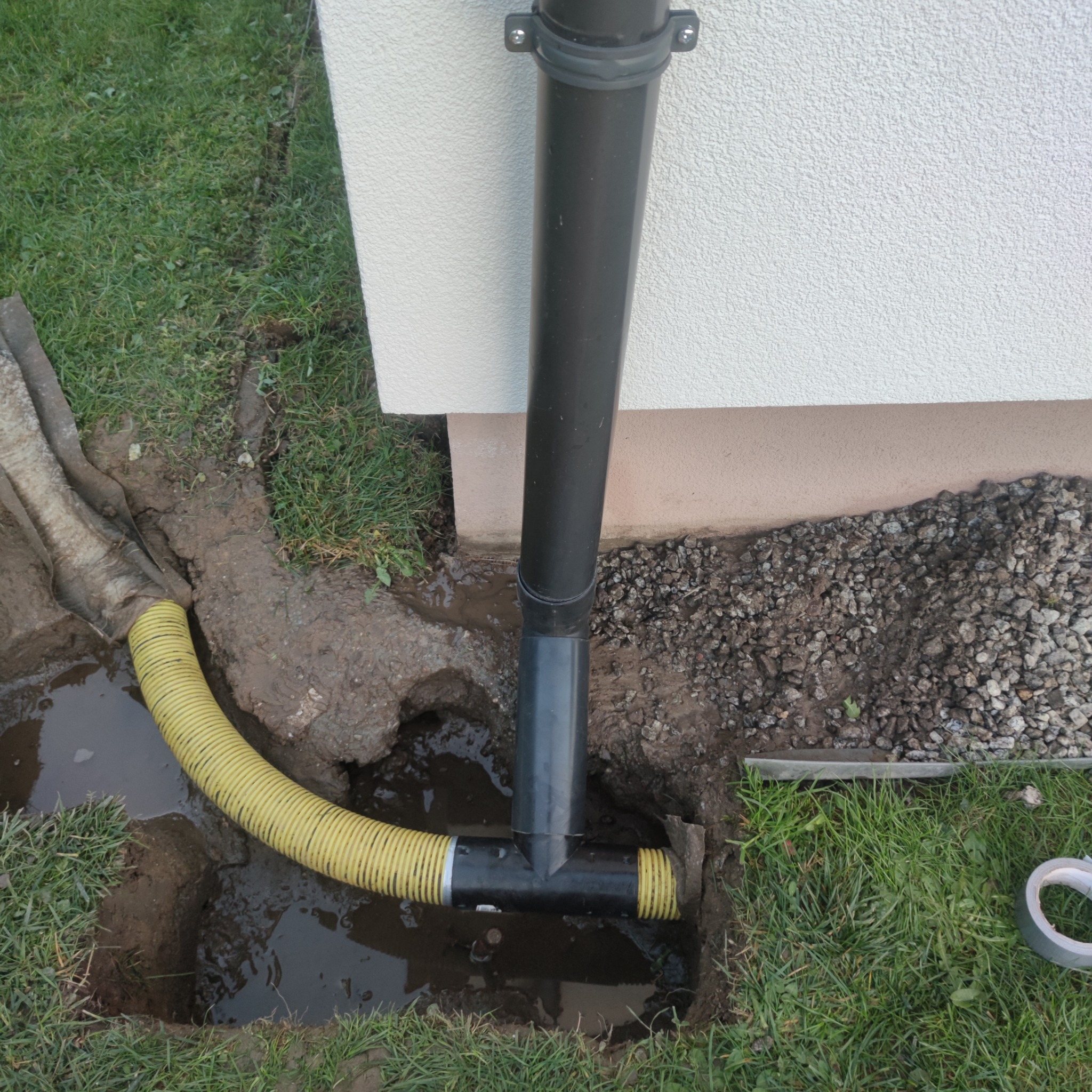 Instalacja odpływu wody deszczowej w wykopie obok budynku z użyciem rury karbowanej i kształtki T, widoczne narzędzia i gruz.