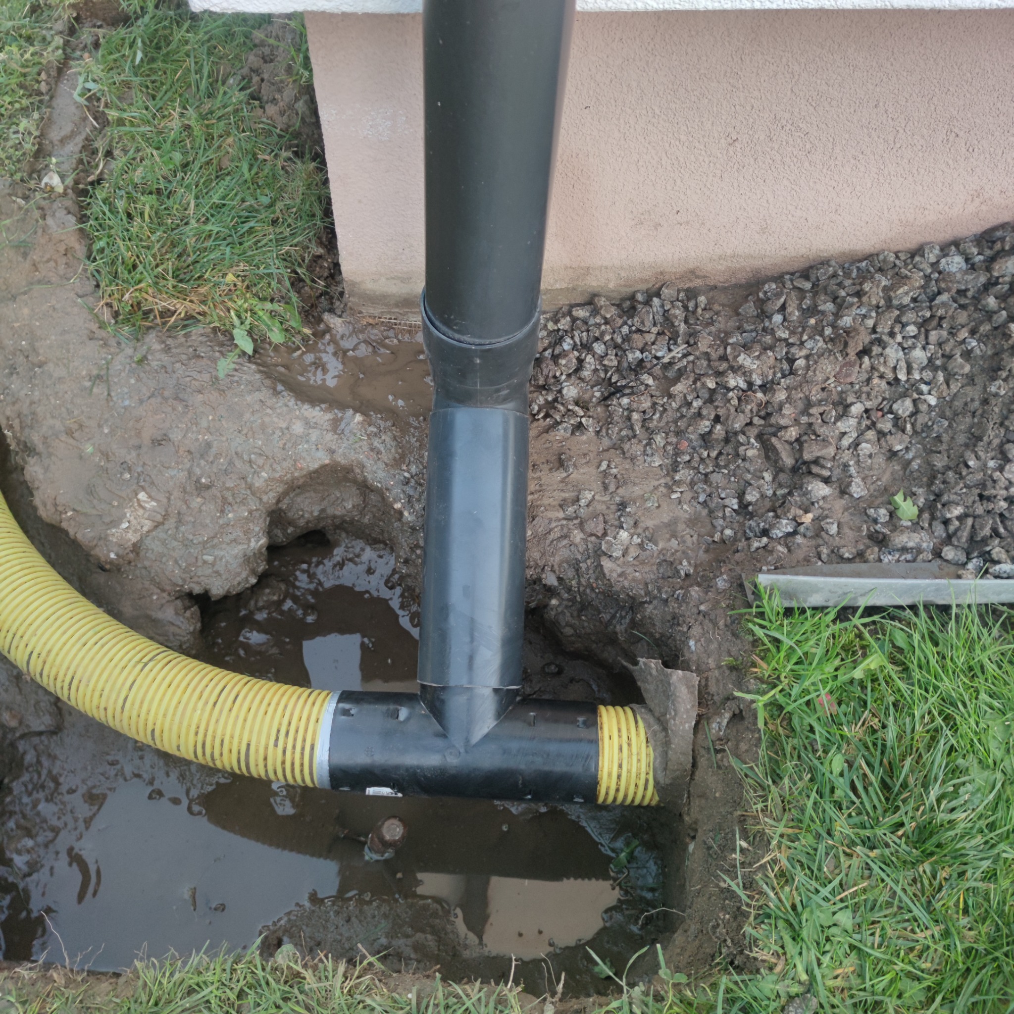 Instalacja odpływu rury spustowej z żółtą rurą drenażową w wykopie przy ścianie domu, widoczne błoto i woda.