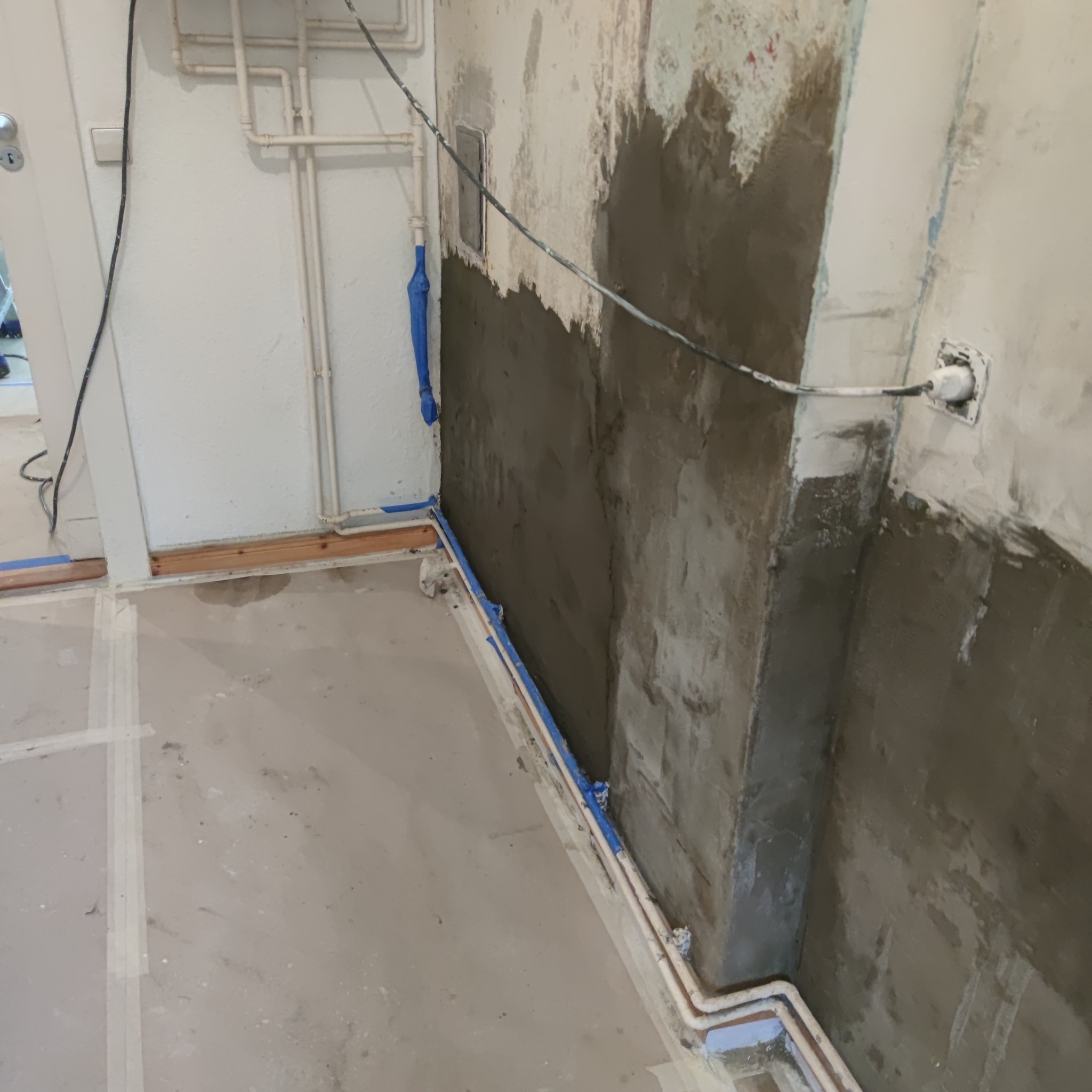 Ściana w trakcie nakładania szarej masy szpachlowej, zabezpieczona niebieską taśmą malarską przy podłodze i rurach, widoczne instalacje wodne i elektryczne
