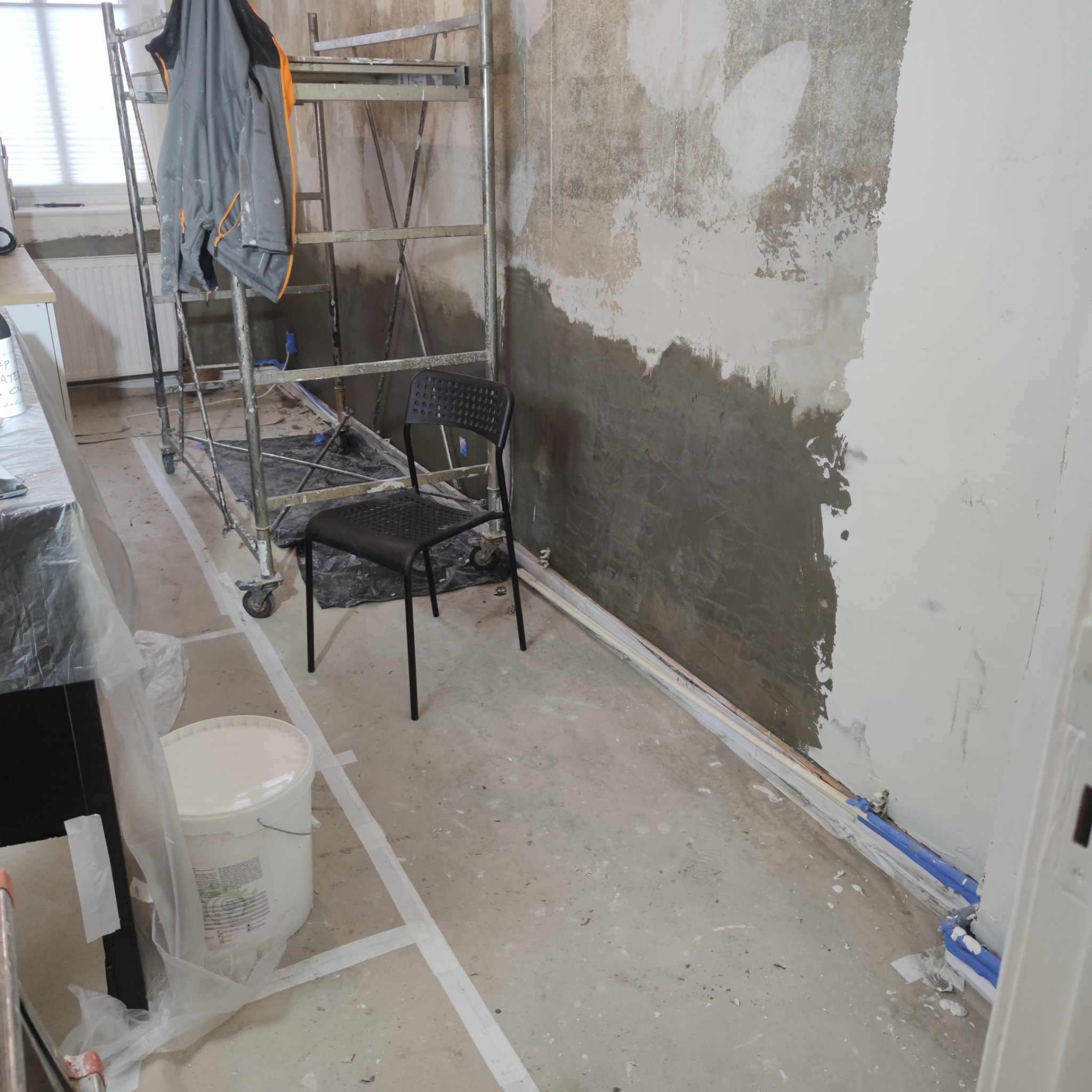 Ściana w trakcie prac przygotowawczych z częściowo nałożoną szarą warstwą, obok metalowe rusztowanie z powieszoną kurtką roboczą, na podłodze biała taśma wyznaczająca obszar roboczy i wiadro z farbą.