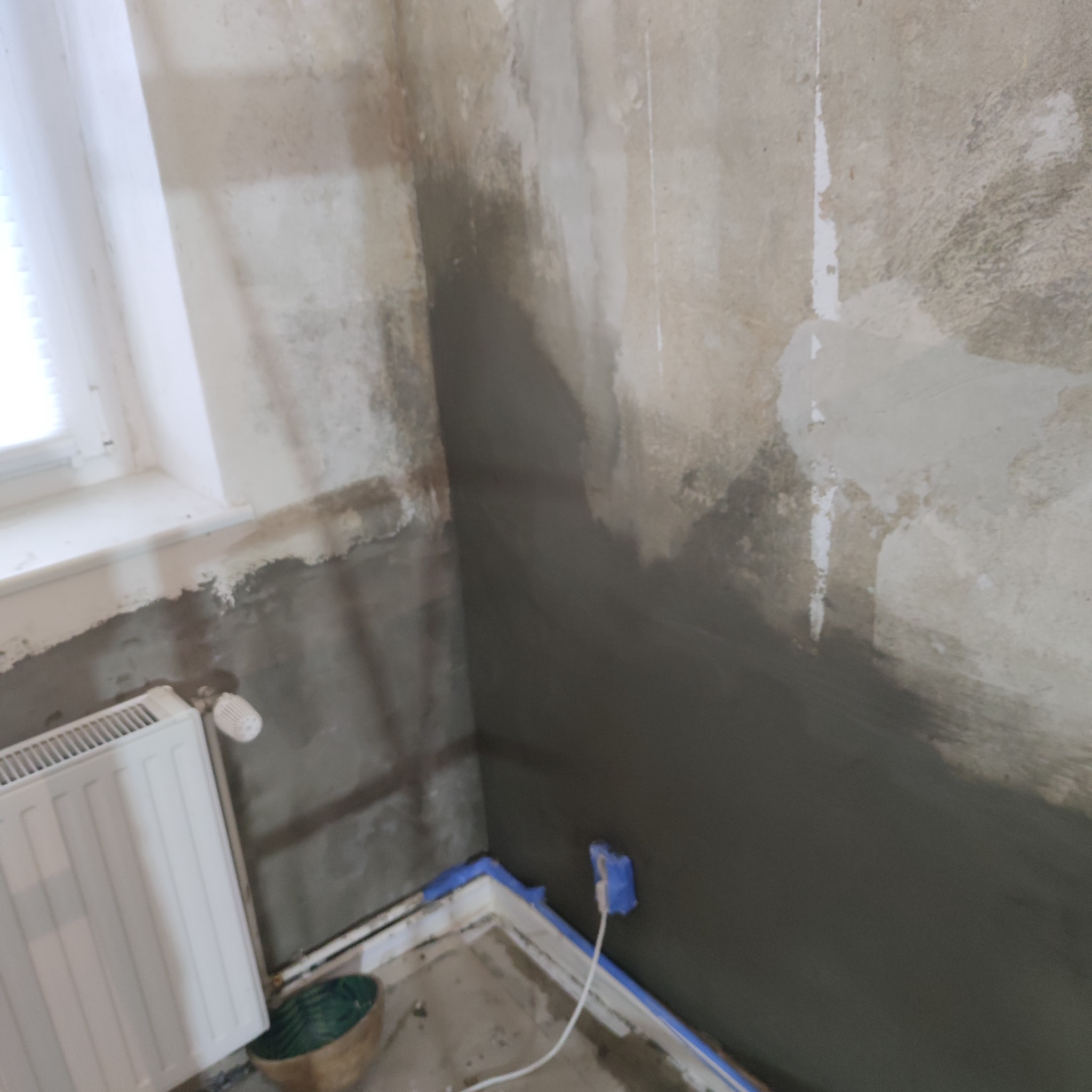 Wnętrze pokoju w trakcie prac przygotowawczych do malowania, ściany częściowo pokryte szarą masą, zabezpieczone taśmą narożniki, widoczny grzejnik i fragment okna z żaluzjami.