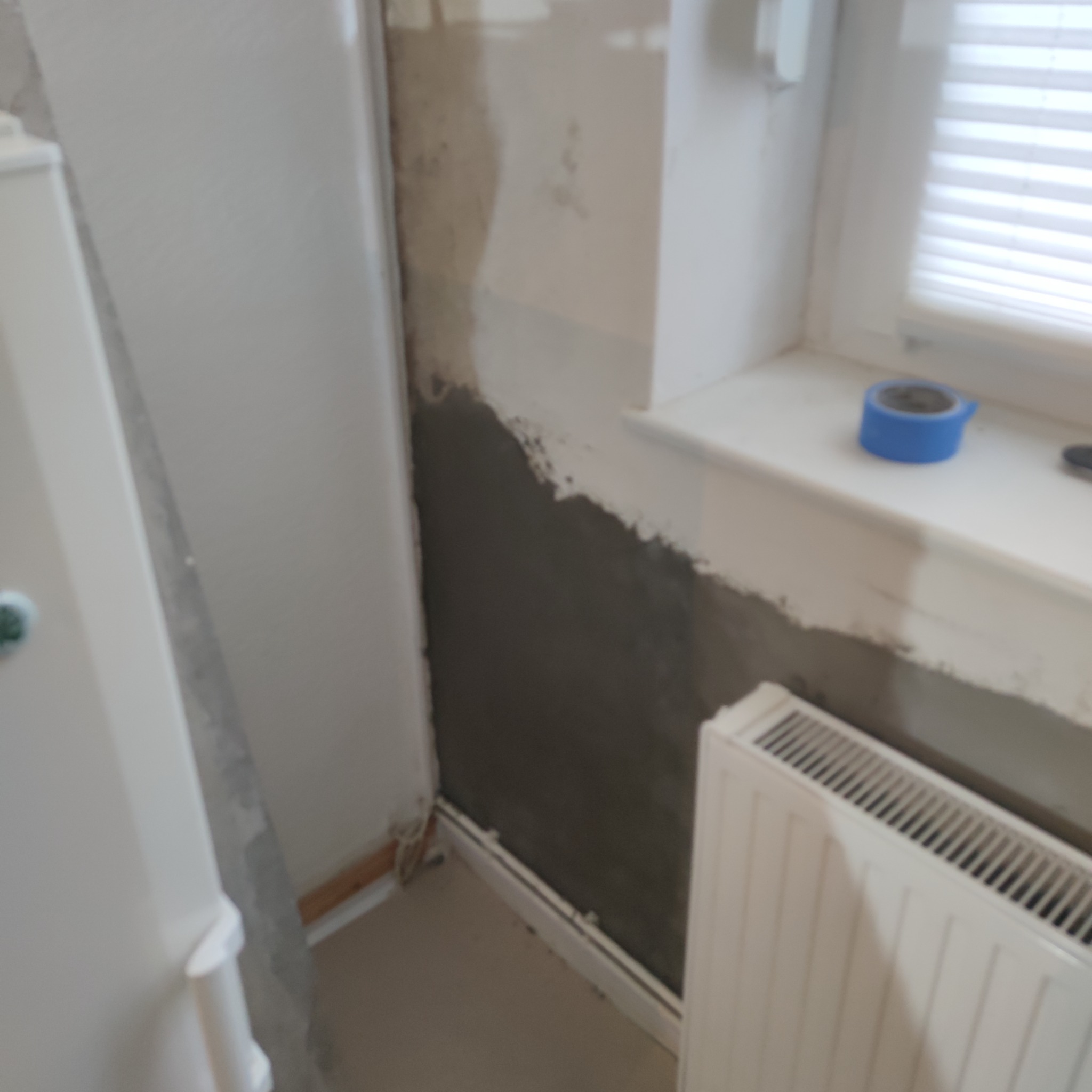 Świeżo nałożona, ciemnoszara warstwa wyrównująca na ścianie w rogu pokoju, widoczny fragment grzejnika i okna z żaluzjami, niebieska taśma malarska na parapecie.