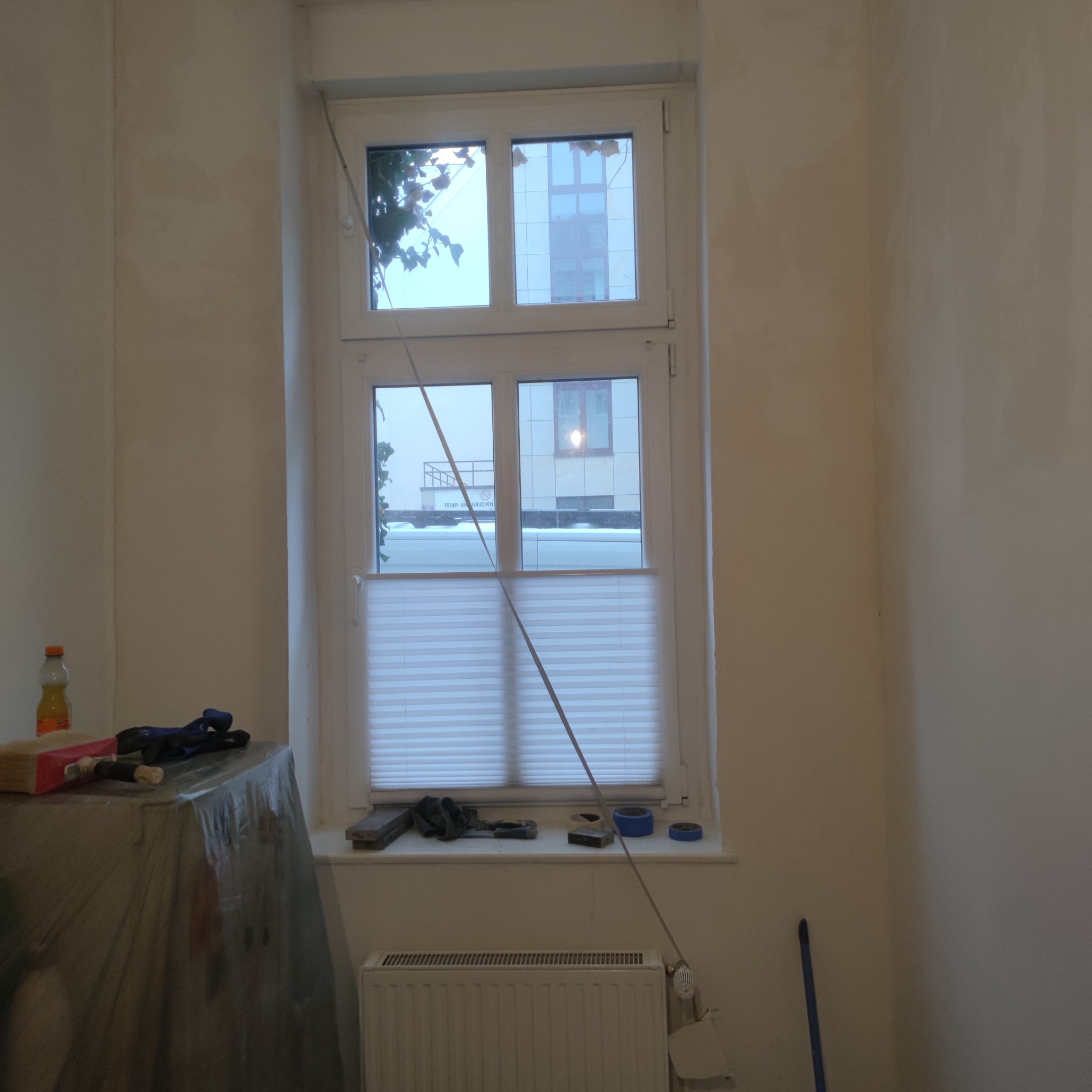 Pokój w trakcie przygotowania do prac wykończeniowych: ściany wstępnie wyrównane, okno z roletą, grzejnik, narzędzia malarskie na zabezpieczonym folią meblu.