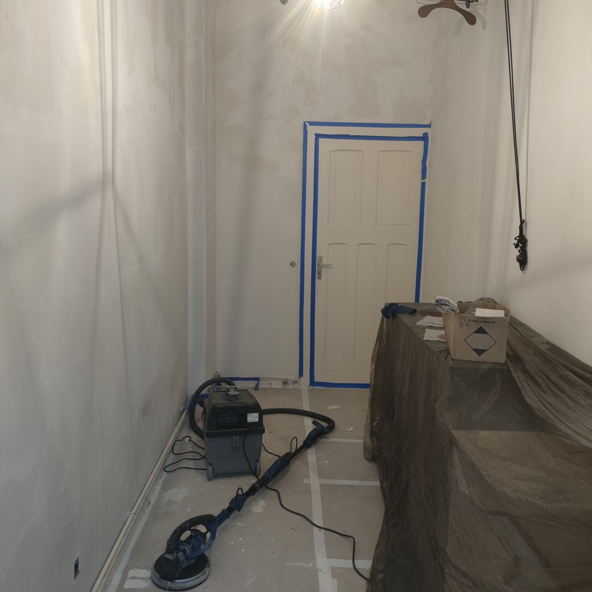 Pokój w trakcie prac: ściany po nałożeniu gładzi, szlifierka do gładzi z odkurzaczem, drzwi zabezpieczone taśmą malarską, podłoga zarysowana siatką linii pomocniczych.