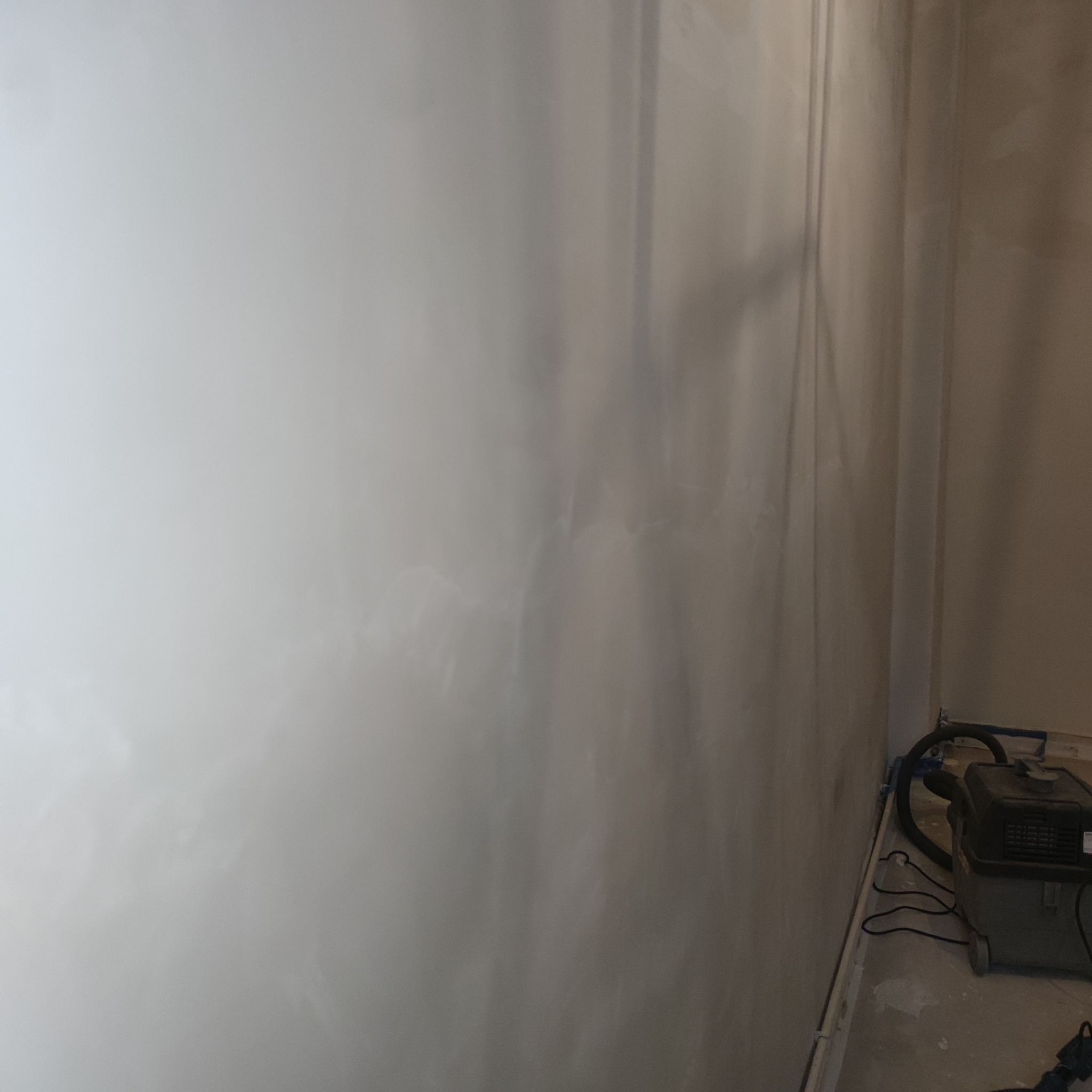 Świeżo nałożona, wilgotna gładź na ścianie, widoczne delikatne smugi i nierówności, w tle odkurzacz przemysłowy.