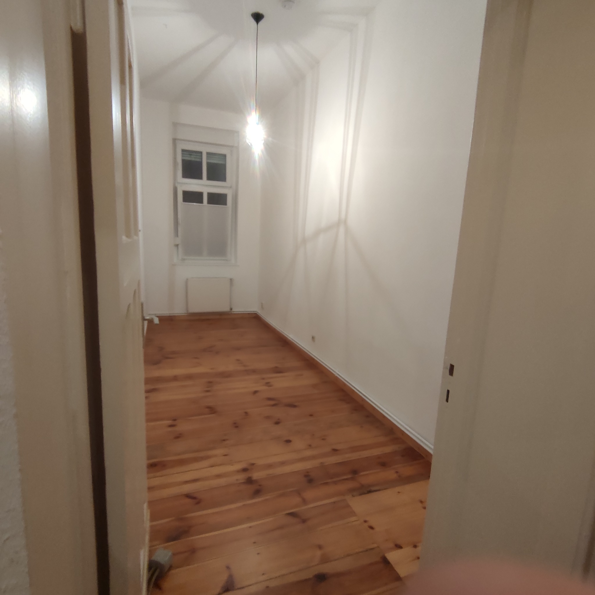 Pusty pokój z drewnianą podłogą i białymi ścianami widziany zza otwartych drzwi, z widocznym oknem i grzejnikiem.