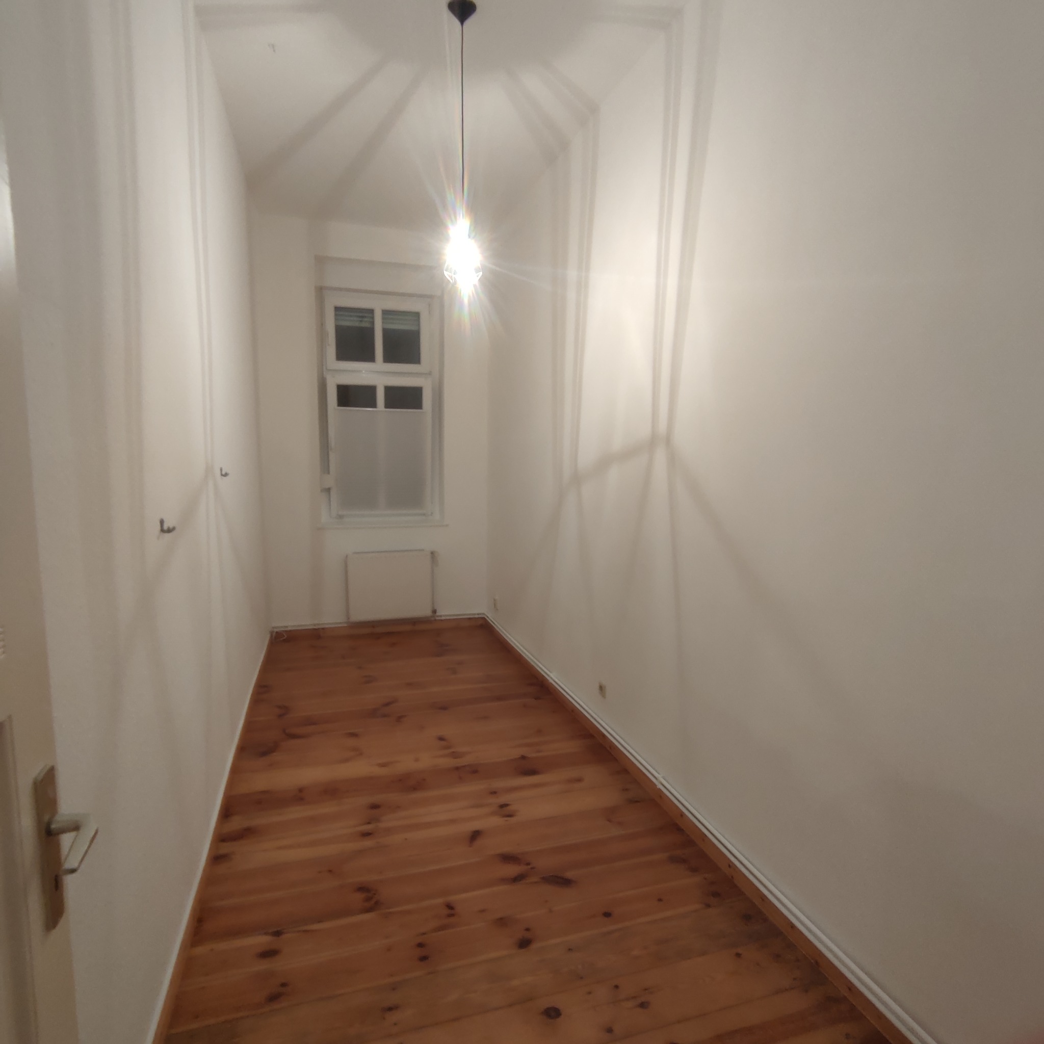 Pusty pokój po remoncie z drewnianą podłogą, białymi ścianami i oknem z matową folią, widoczne haczyki na ścianie i żarówka sufitowa.