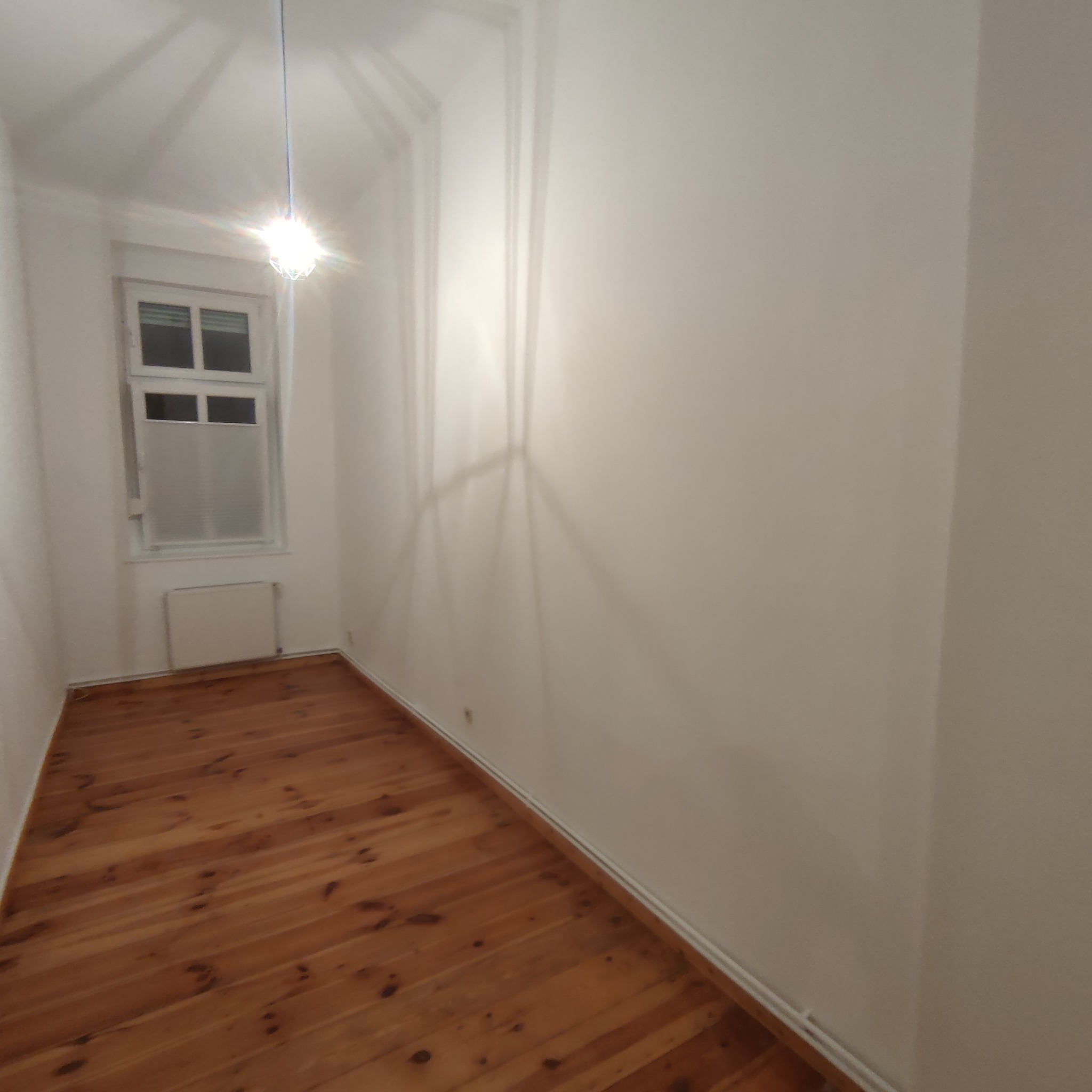 Pusty pokój po odświeżeniu, widoczne drewniane podłogi, białe ściany i okno z grzejnikiem, oświetlony przez wiszącą lampę z żarówką.