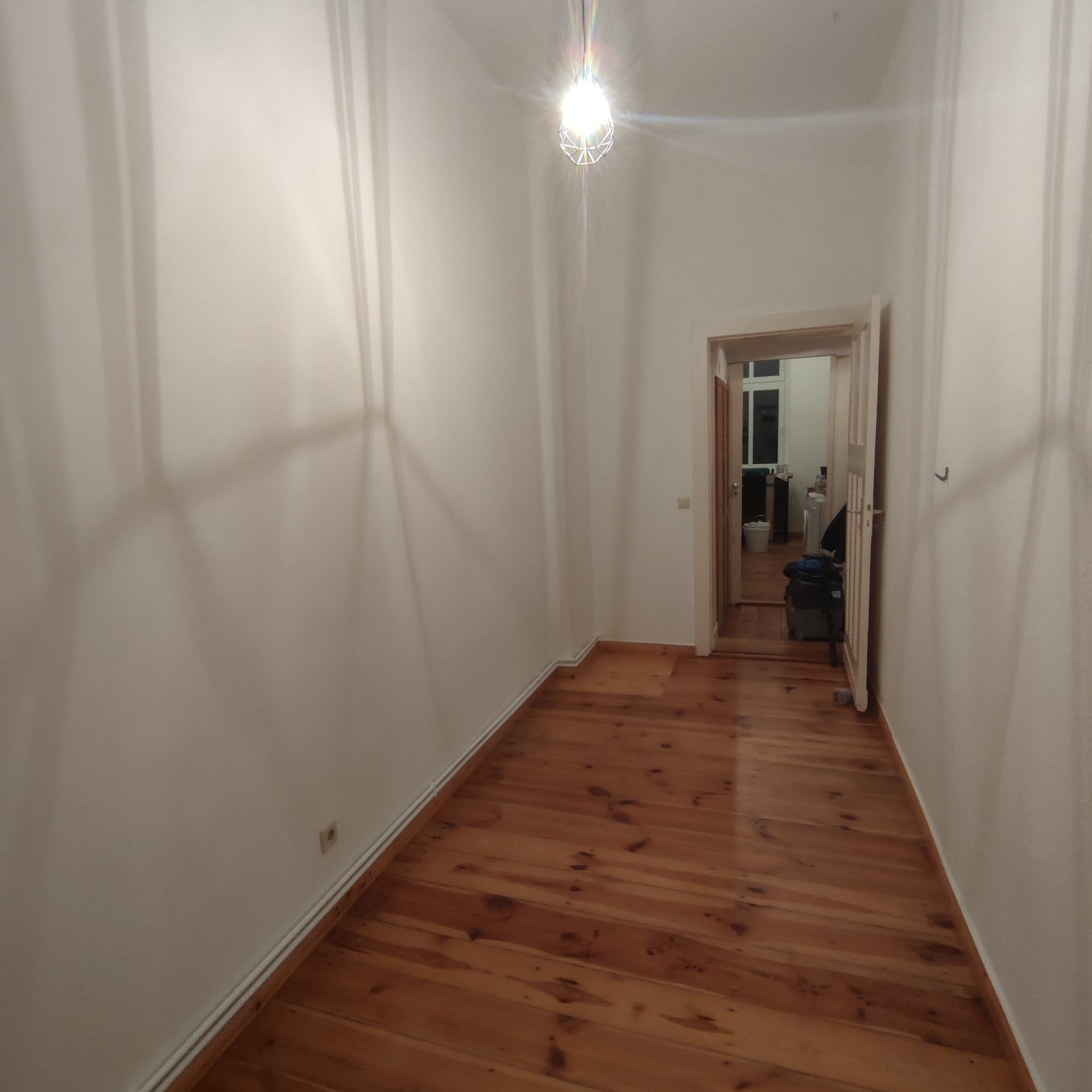 Puste, świeżo odnowione pomieszczenie z drewnianą podłogą i białymi ścianami, widok na sąsiedni pokój przez otwarte drzwi, żyrandol w geometrycznej oprawie na suficie.