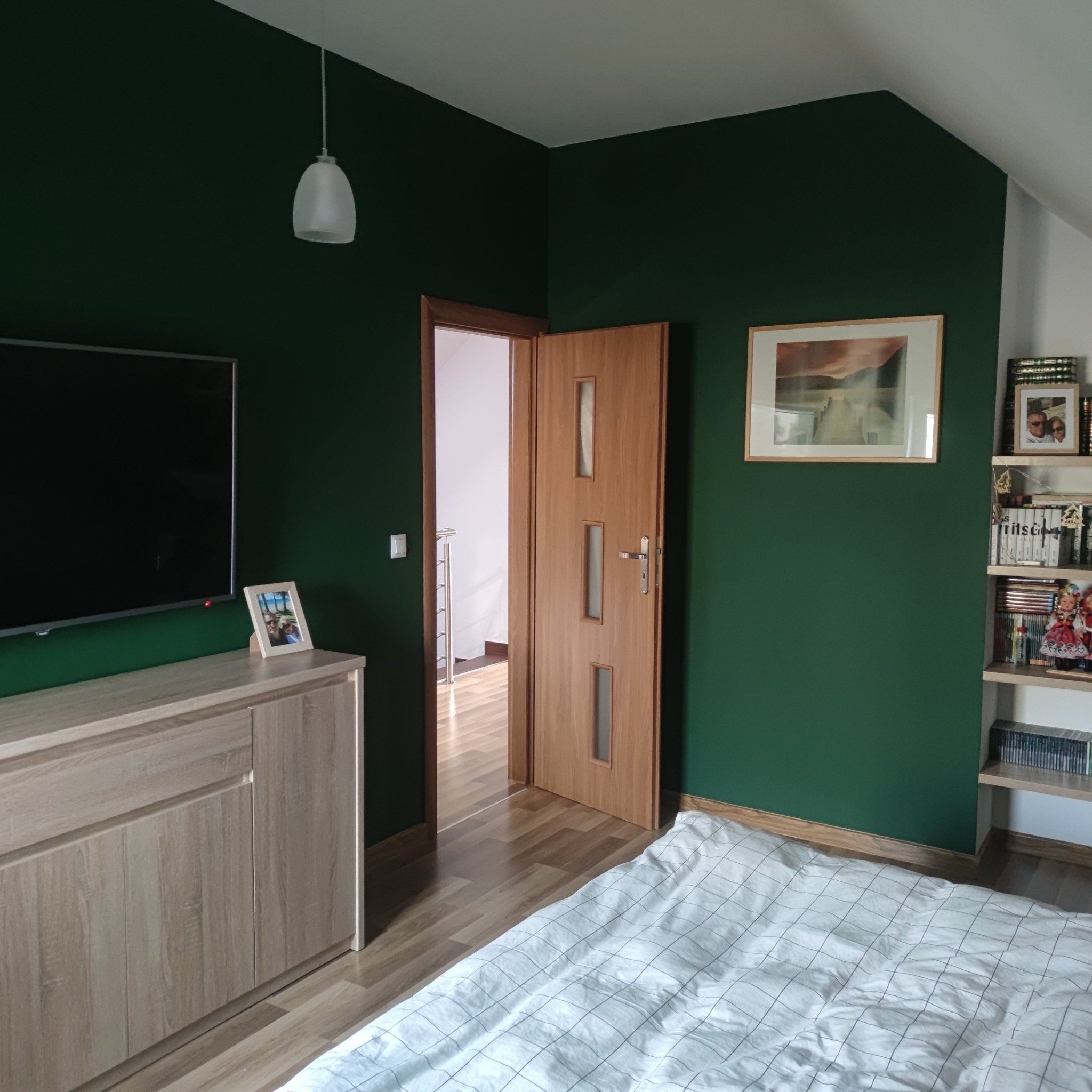 Sypialnia na poddaszu z ciemnozielonymi ścianami, jasną komodą pod telewizorem, drewnianymi drzwiami i łóżkiem w kratę.