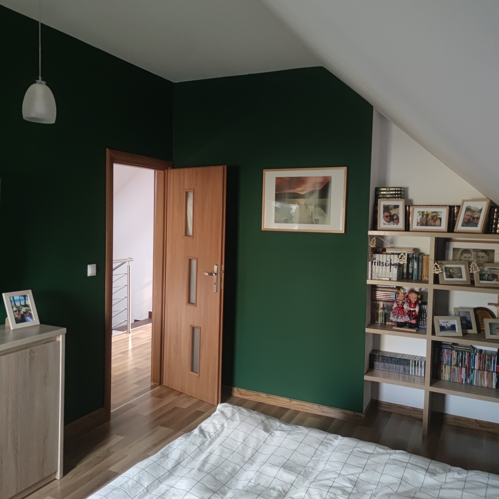 Pokój na poddaszu z ciemnozielonymi ścianami, drewnianą podłogą i regałem z dekoracjami, oświetlony wiszącą lampą.