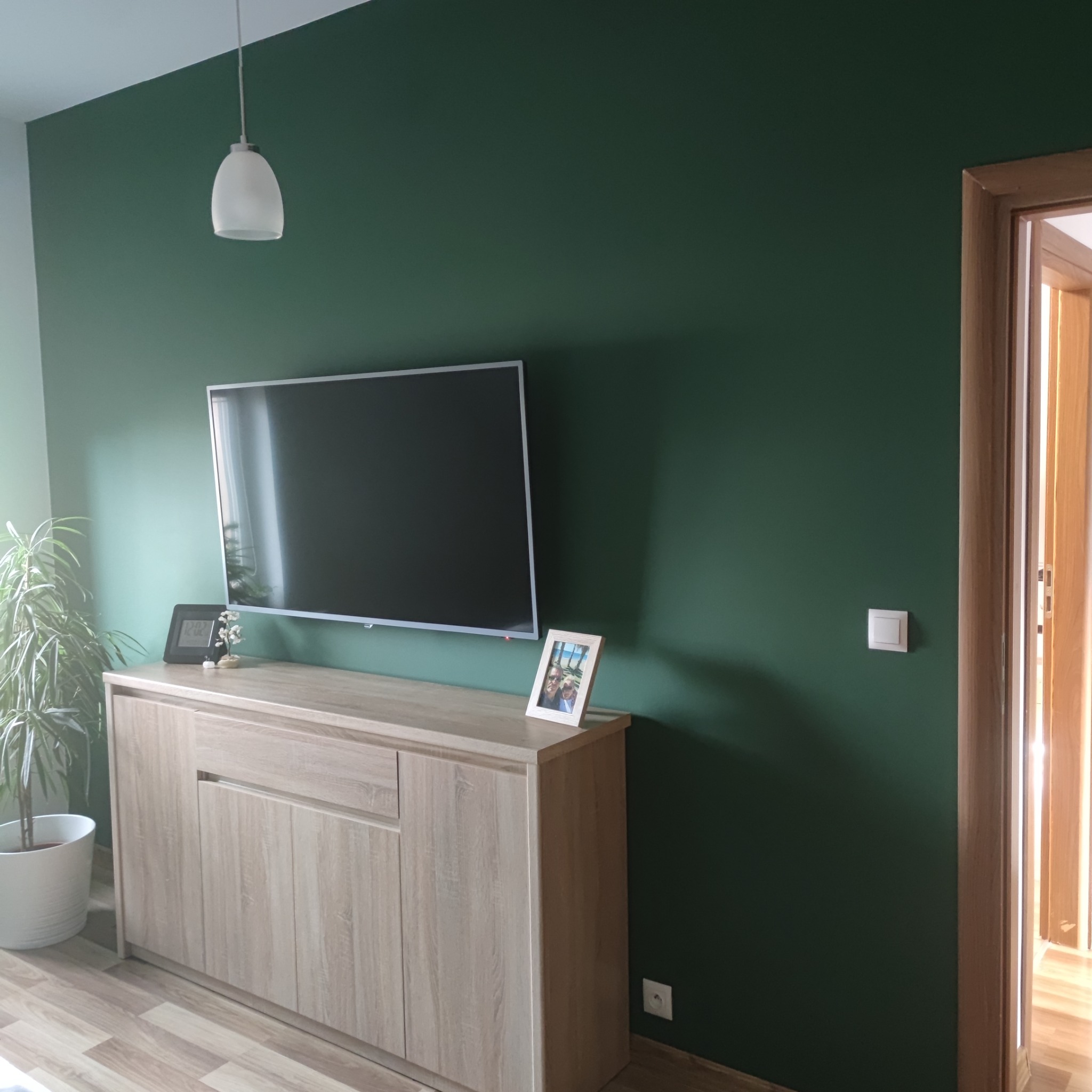 Pokój z ciemnozieloną ścianą, jasną komodą pod telewizorem, na której stoją ramki ze zdjęciami i zegar, oraz rośliną w białej doniczce.