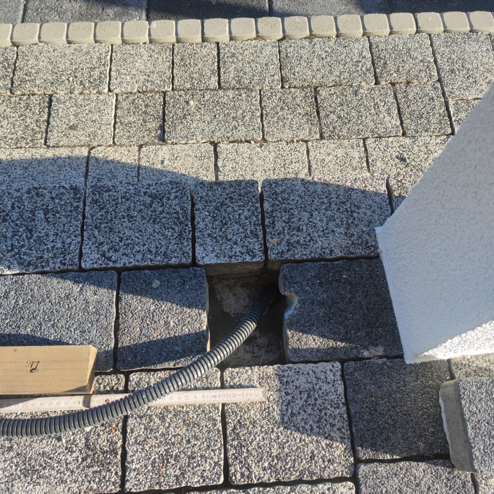 Układanie szarej kostki brukowej z widocznym kablem w peszlu, częściowo odsłoniętym, oraz miarką stolarską