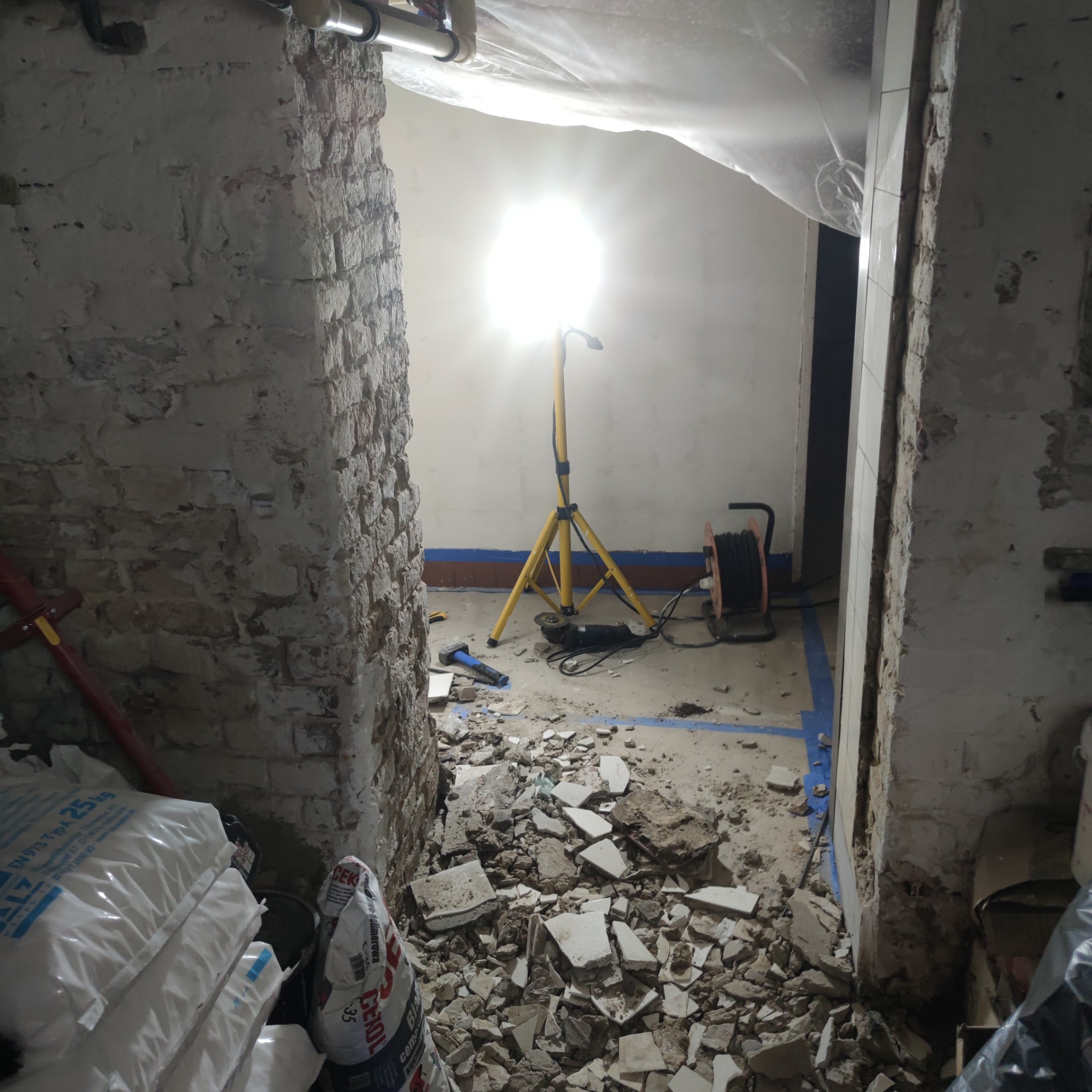 Demontaż płytek w pomieszczeniu z ceglanymi ścianami, widoczne gruz, worki z zaprawą, szlifierka kątowa i lampa budowlana na trójnogu.