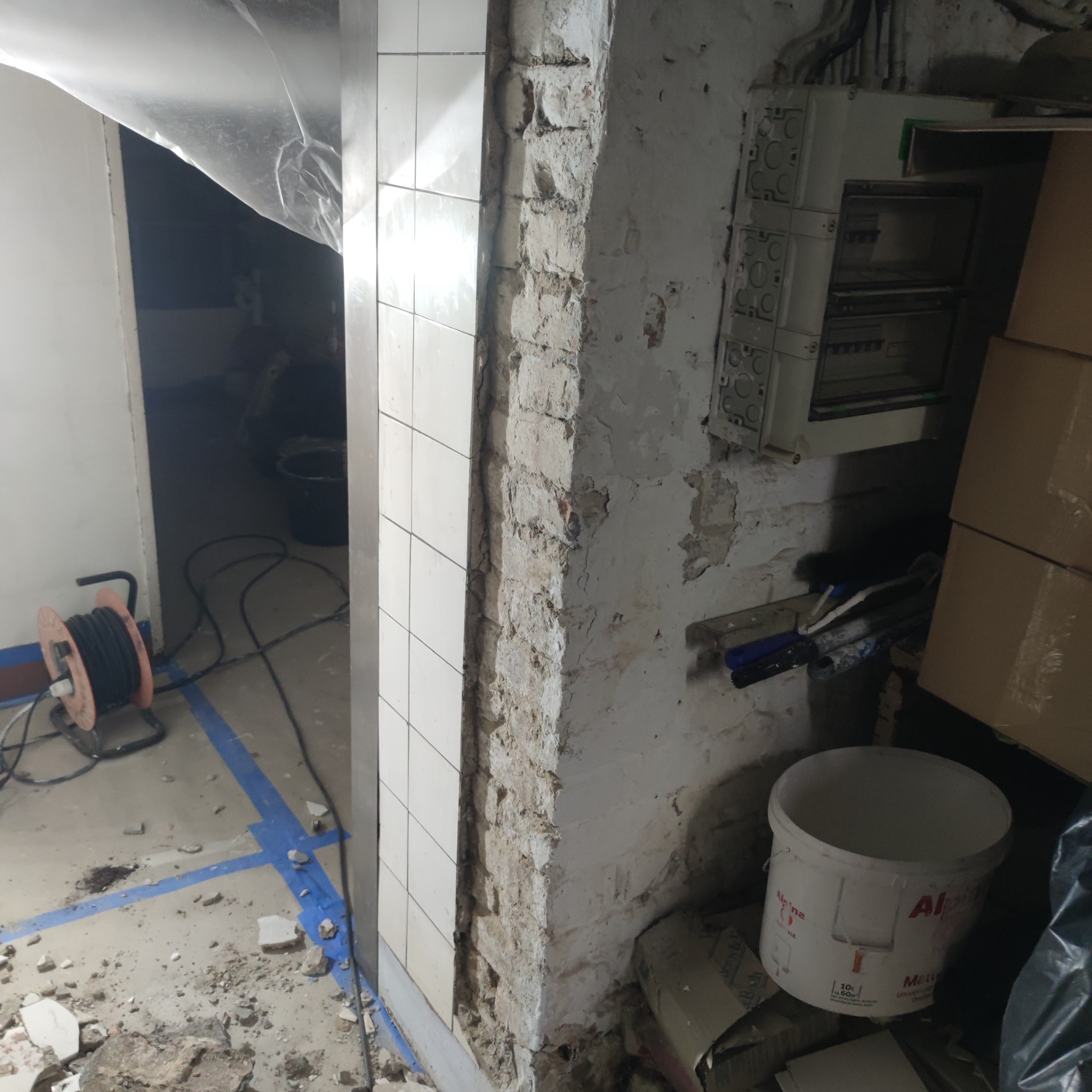 Demontaż płytek i odsłonięta surowa cegła w trakcie prac remontowych, widoczne kable elektryczne, puszki i wiadro farby.