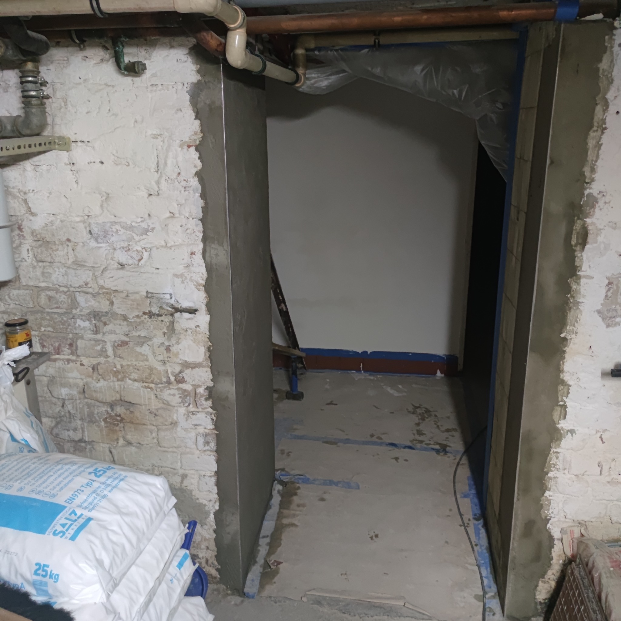 Przygotowanie do prac wykończeniowych: ościeżnica drzwiowa po osadzeniu, obłożona świeżą warstwą zaprawy, widoczne worki z materiałami budowlanymi i surowe cegły w tle.