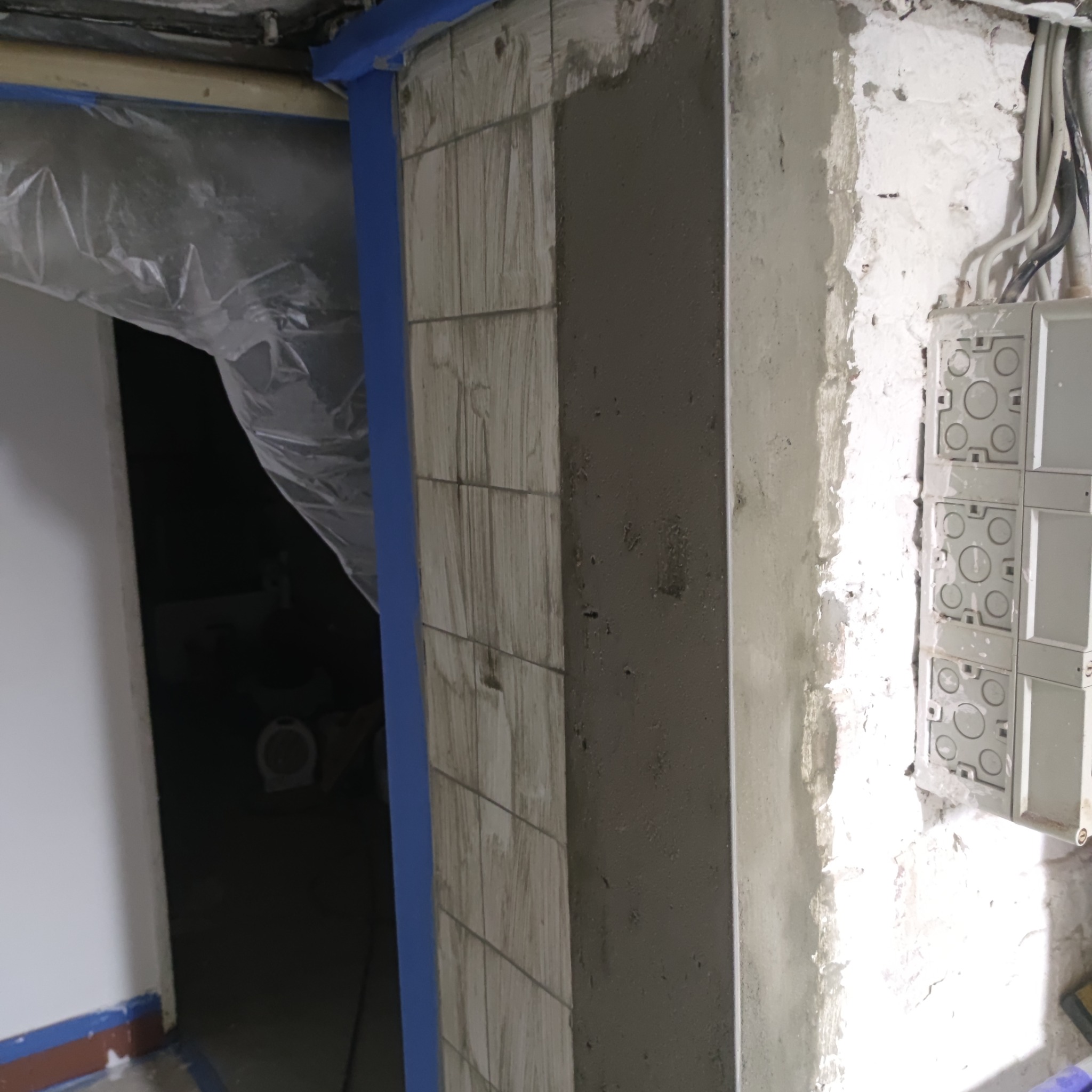 Narożnik ściany w trakcie prac: częściowo położone płytki imitujące drewno zabezpieczone niebieską taśmą, obok fragment ściany pokryty świeżą warstwą szarej masy, widoczne przewody i puszki...