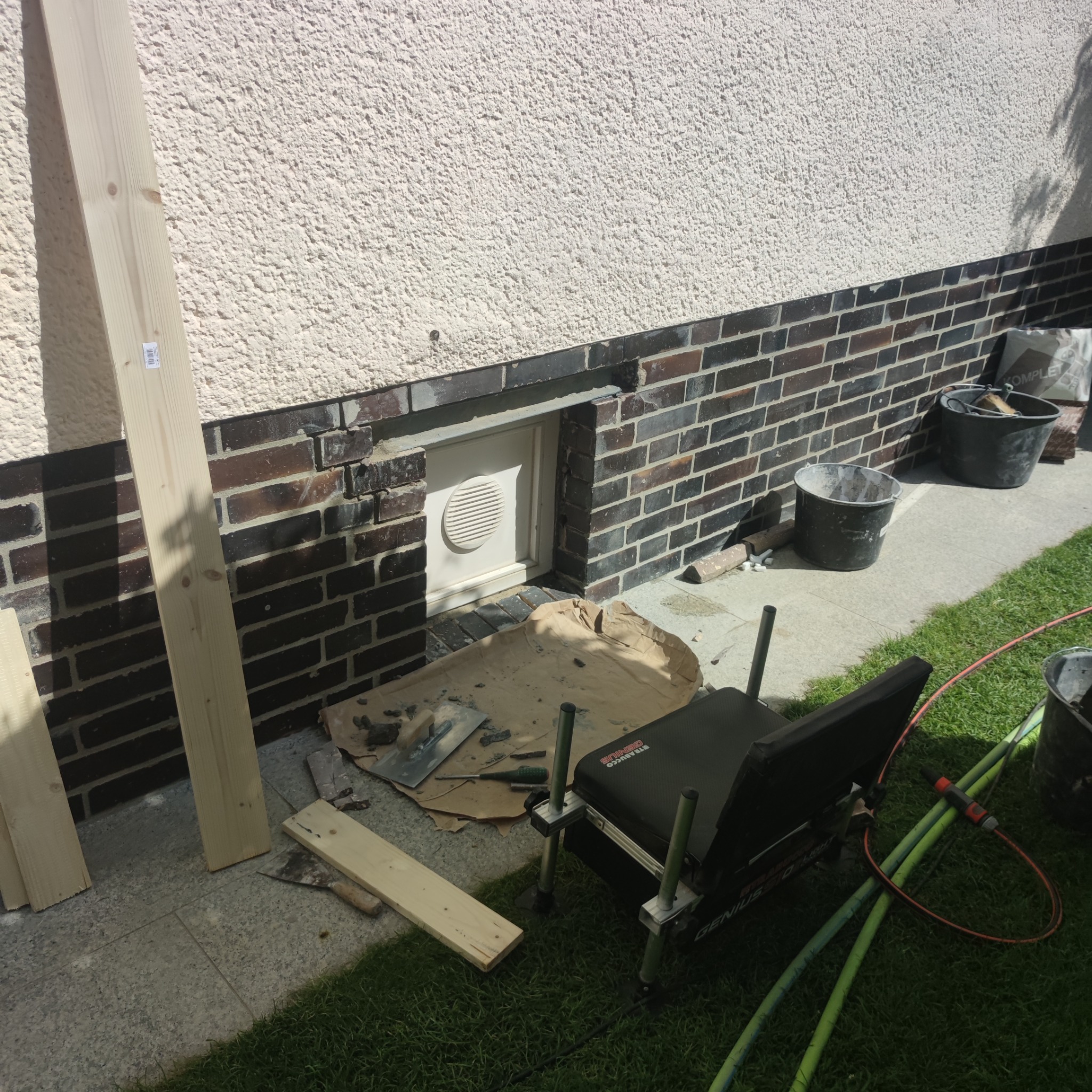 Przygotowania do prac murarskich przy elewacji budynku: deski, kielnie i wiaderka ustawione obok ściany z ceglaną podmurówką i wentylatorem.