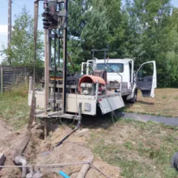 Wiercenie studni na działce: widoczna wiertnica zamontowana na samochodzie dostawczym, otwór w ziemi z wypływającą wodą, elementy instalacji wodnej i narzędzia. W tle drzewa i słup elektryczny.