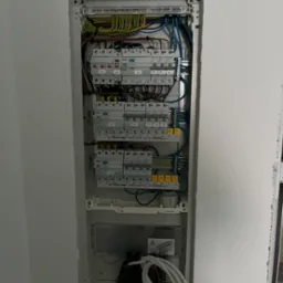 Rozdzielnica elektryczna wnękowa, widoczne zabezpieczenia nadprądowe, różnicowoprądowe, przewody w kolorach brązowym, niebieskim, żółto-zielonym, oraz przewód zasilający zwinięty na dole wnęki.