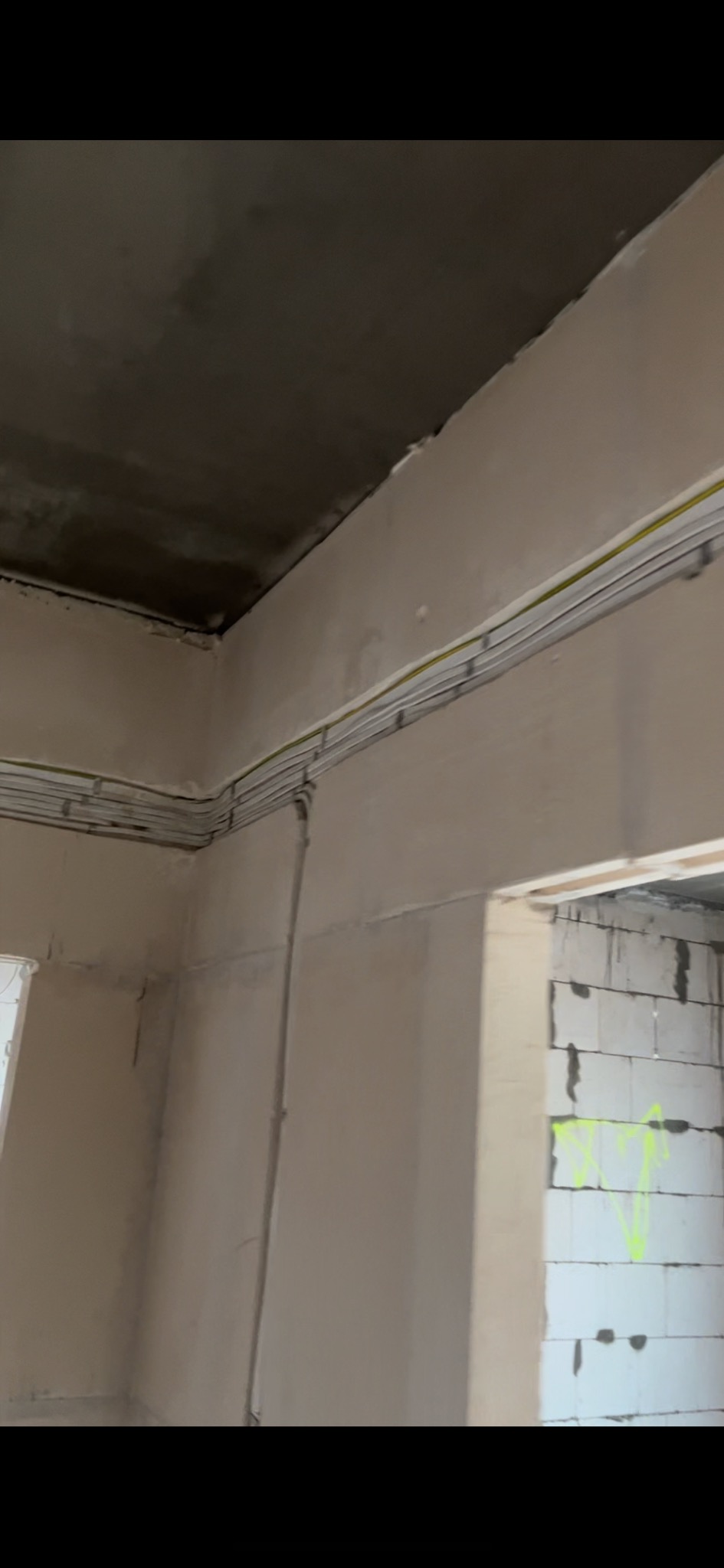 Wnętrze w stanie surowym z widocznymi przewodami elektrycznymi poprowadzonymi pod sufitem i wzdłuż ściany, widok na mur z białych bloczków z zielonymi oznaczeniami.