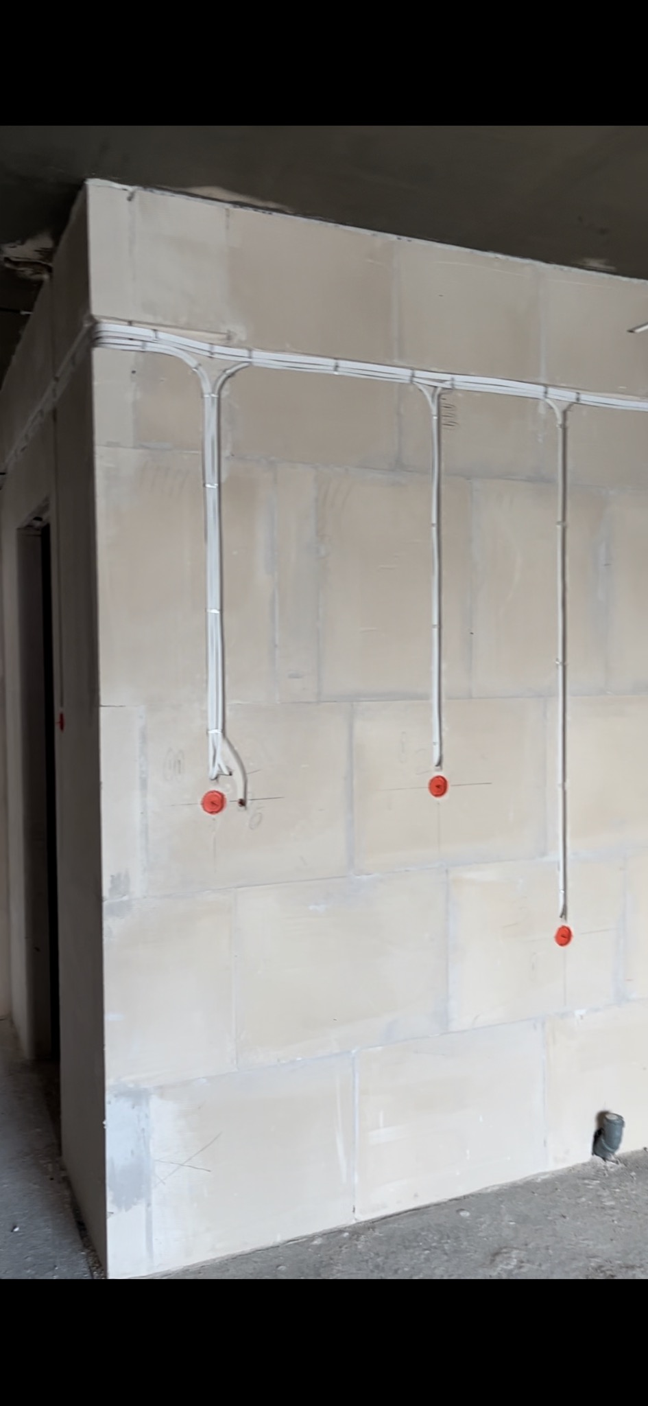 Białe kable elektryczne prowadzone po ścianie z bloków betonowych, z widocznymi puszkami instalacyjnymi zabezpieczonymi czerwonymi zaślepkami.