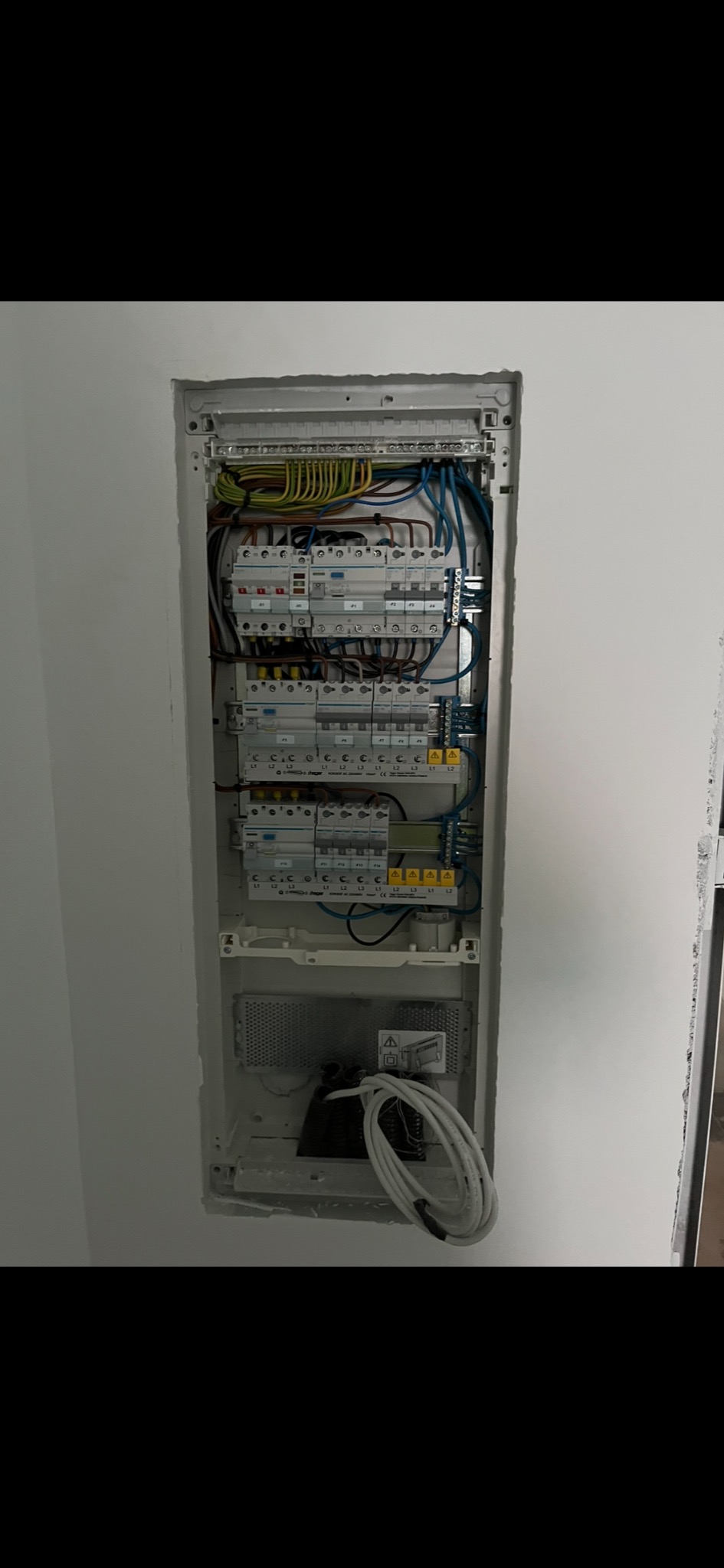 Rozdzielnica elektryczna wnękowa, widoczne zabezpieczenia nadprądowe, różnicowoprądowe, przewody w kolorach brązowym, niebieskim, żółto-zielonym, oraz przewód zasilający zwinięty na dole wnęki.