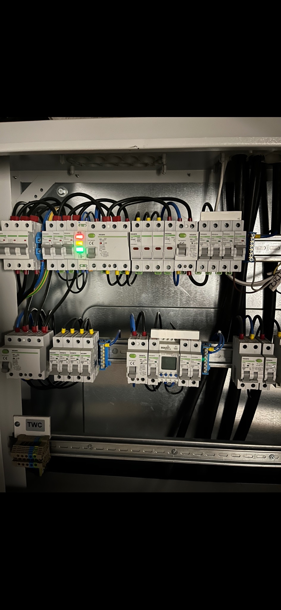 Wnętrze szafy rozdzielczej z zamontowanymi bezpiecznikami, przewodami w kolorach niebieskim, żółtym i czarnym, oraz zieloną diodą sygnalizacyjną włączonego obwodu.
