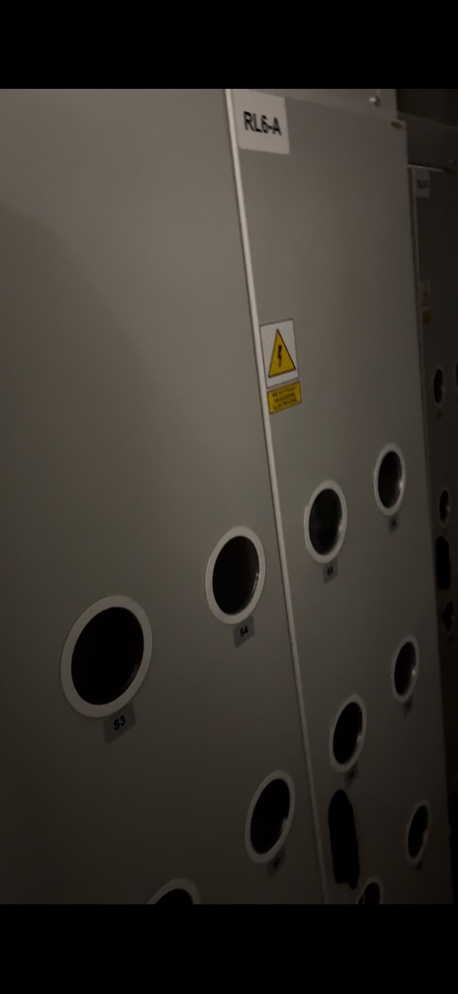 Szara szafa elektryczna RL6-A z okrągłymi otworami oznaczonymi numerami i znakiem ostrzegawczym o wysokim napięciu, widziana pod kątem w słabym oświetleniu.